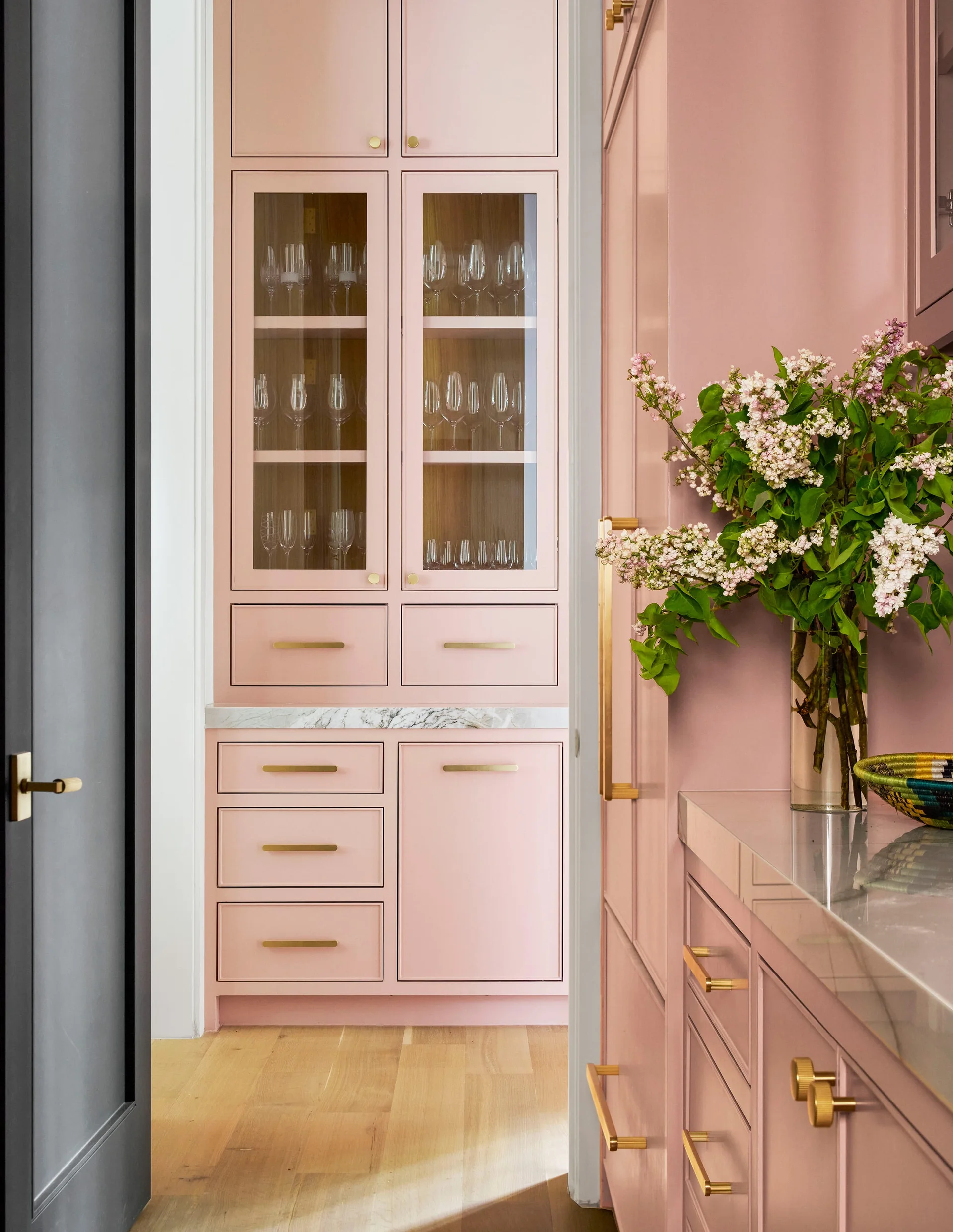 Built-in pink cabinetry in a butler's pantry features glass-front doors showing glassware, a white marble countertop, and brass drawer pulls.