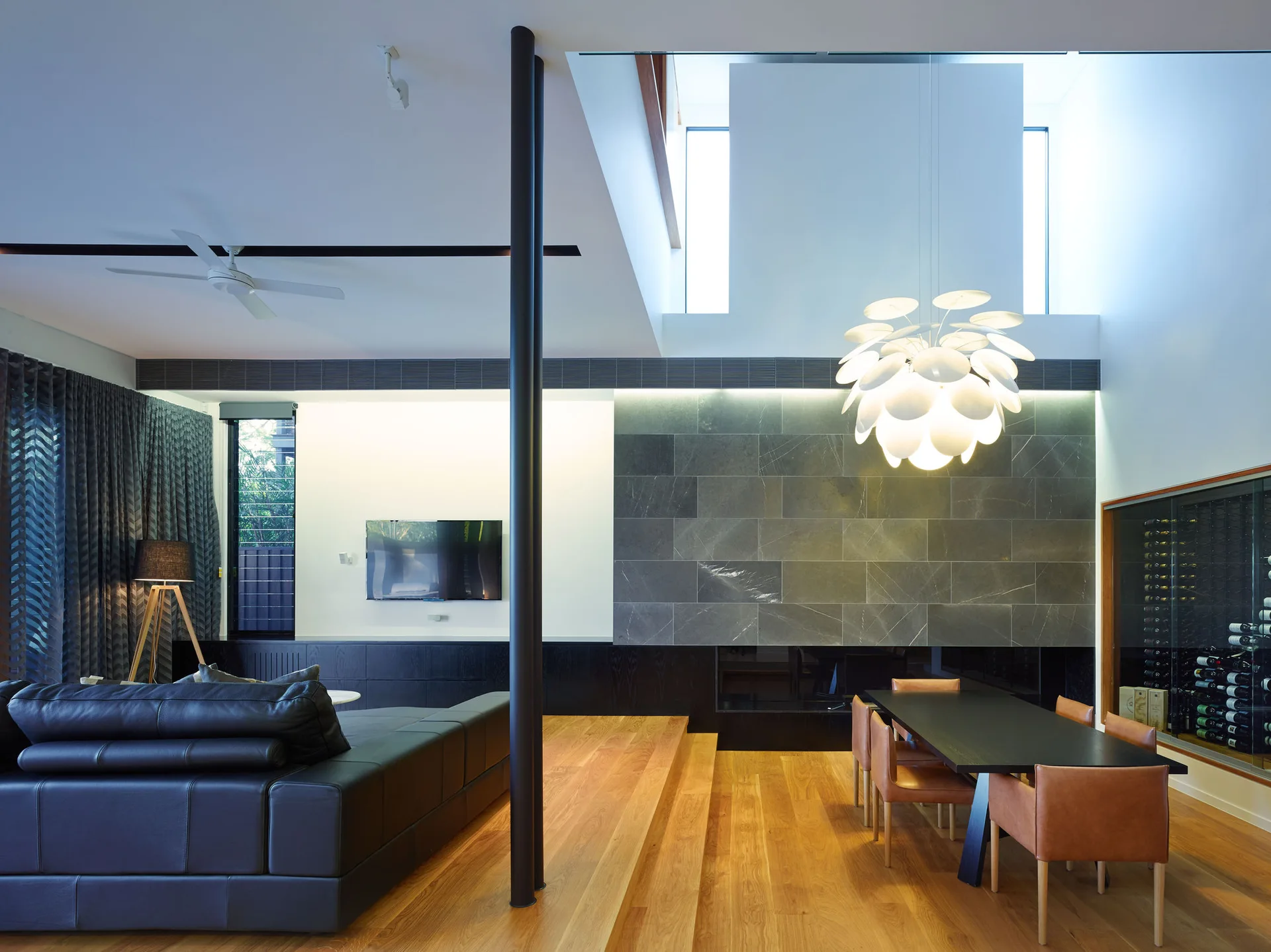 A split-level living area with a black leather sofa is separated by steps from a dining room with tan leather chairs and a stone feature wall.