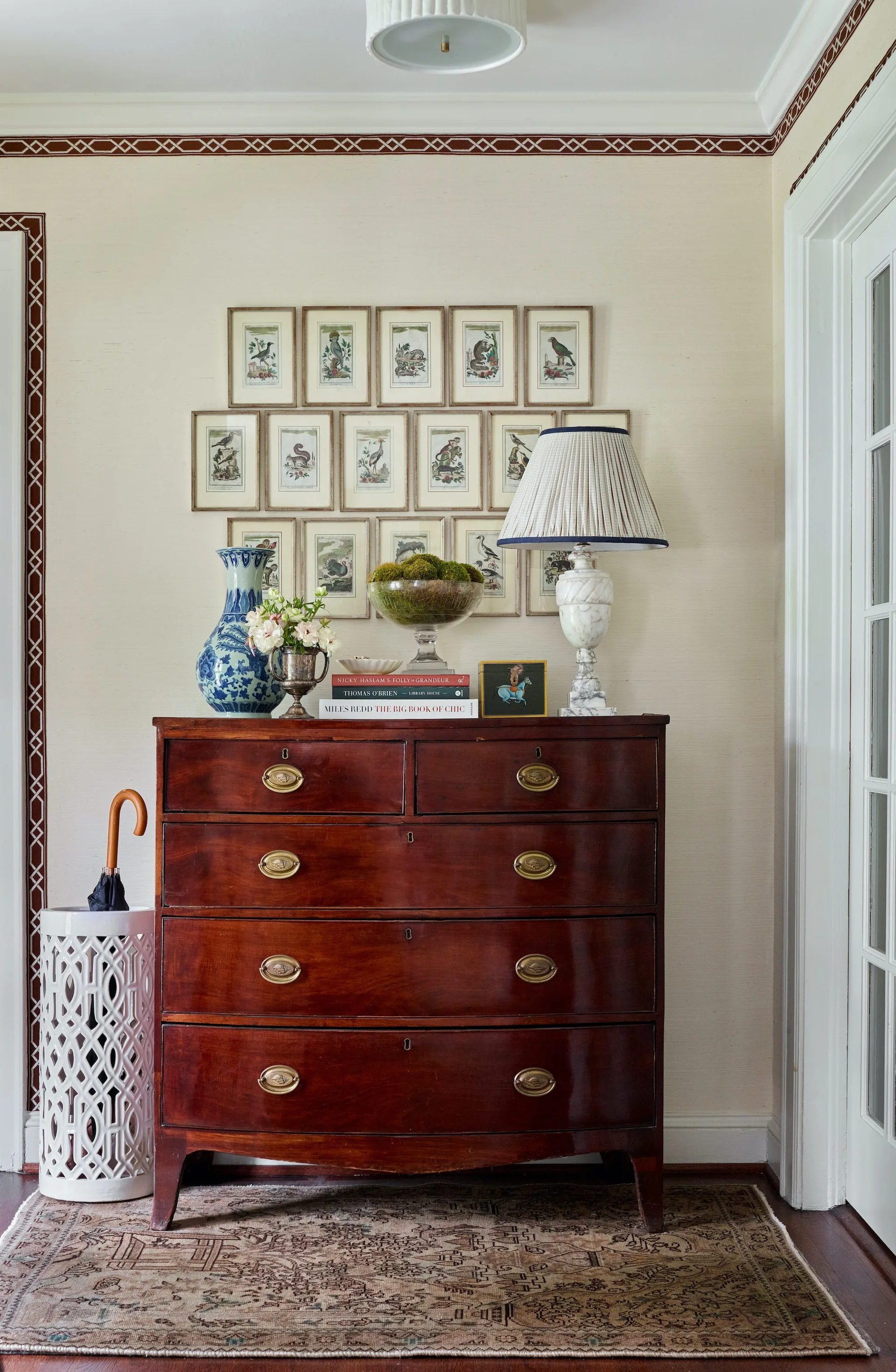 An antique mahogany chest of drawers in an entryway features a ceramic vase, table lamp, and a symmetrical grid of framed botanical prints on the wall.