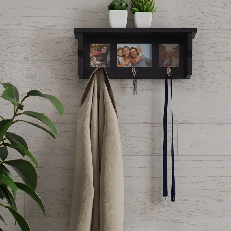 A black wall-mounted shelf featuring three integrated picture frames and hanging hooks is mounted on a light grey wood-plank wall.