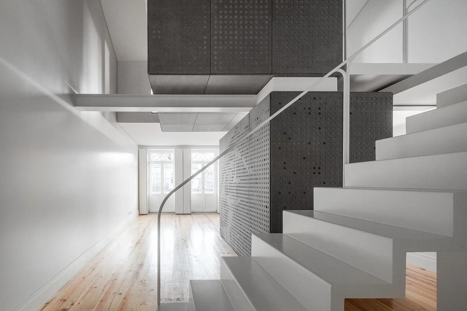 A minimalist white staircase stands beside a dark grey perforated storage unit in a bright room with light wood floors.