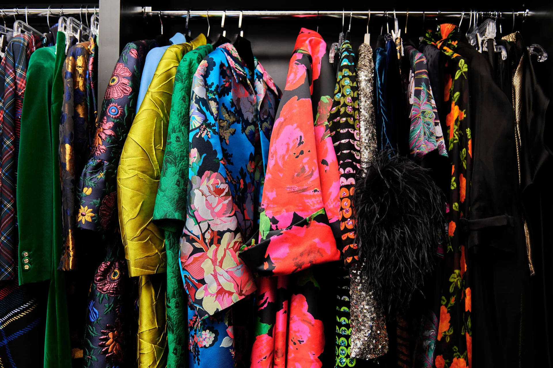 Brightly colored, patterned, and textured garments hang closely together on hangers along a dark closet clothing rack.