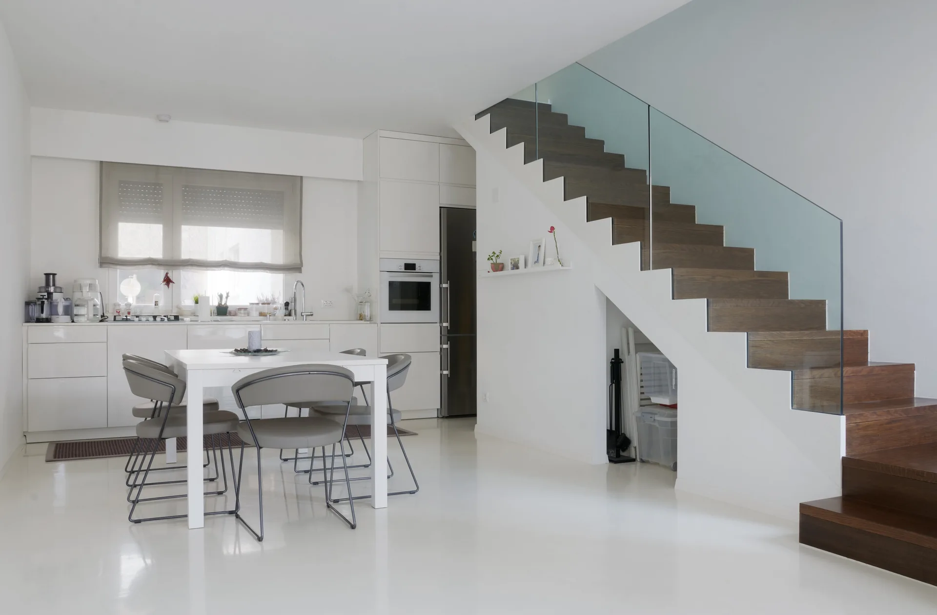 A minimalist white kitchen and dining area features a prominent staircase with dark wooden steps, a glass railing, and grey dining chairs around a white table.