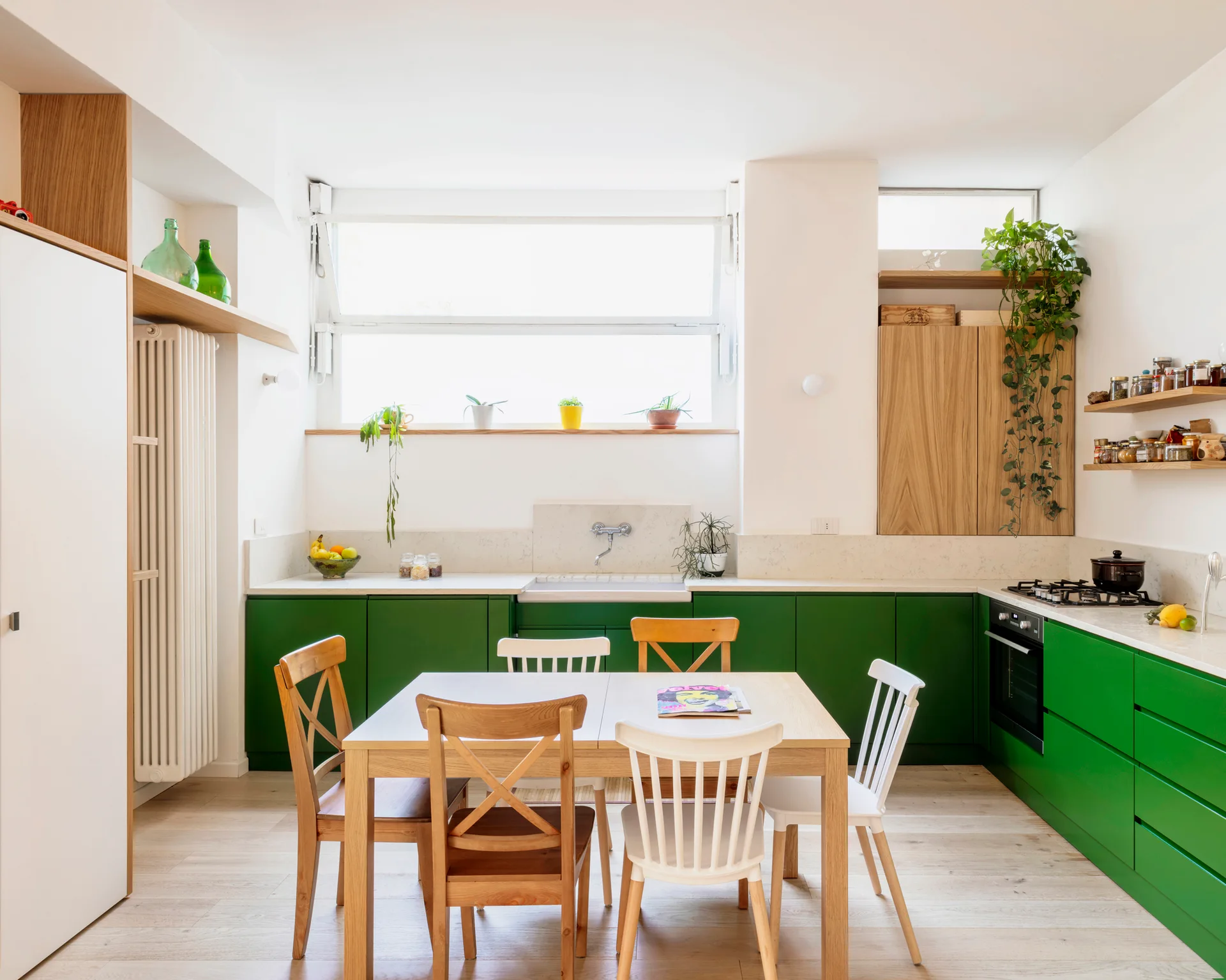 A bright kitchen featuring deep green base cabinets, a light wood dining table with mixed chairs, and a large window above the sink.