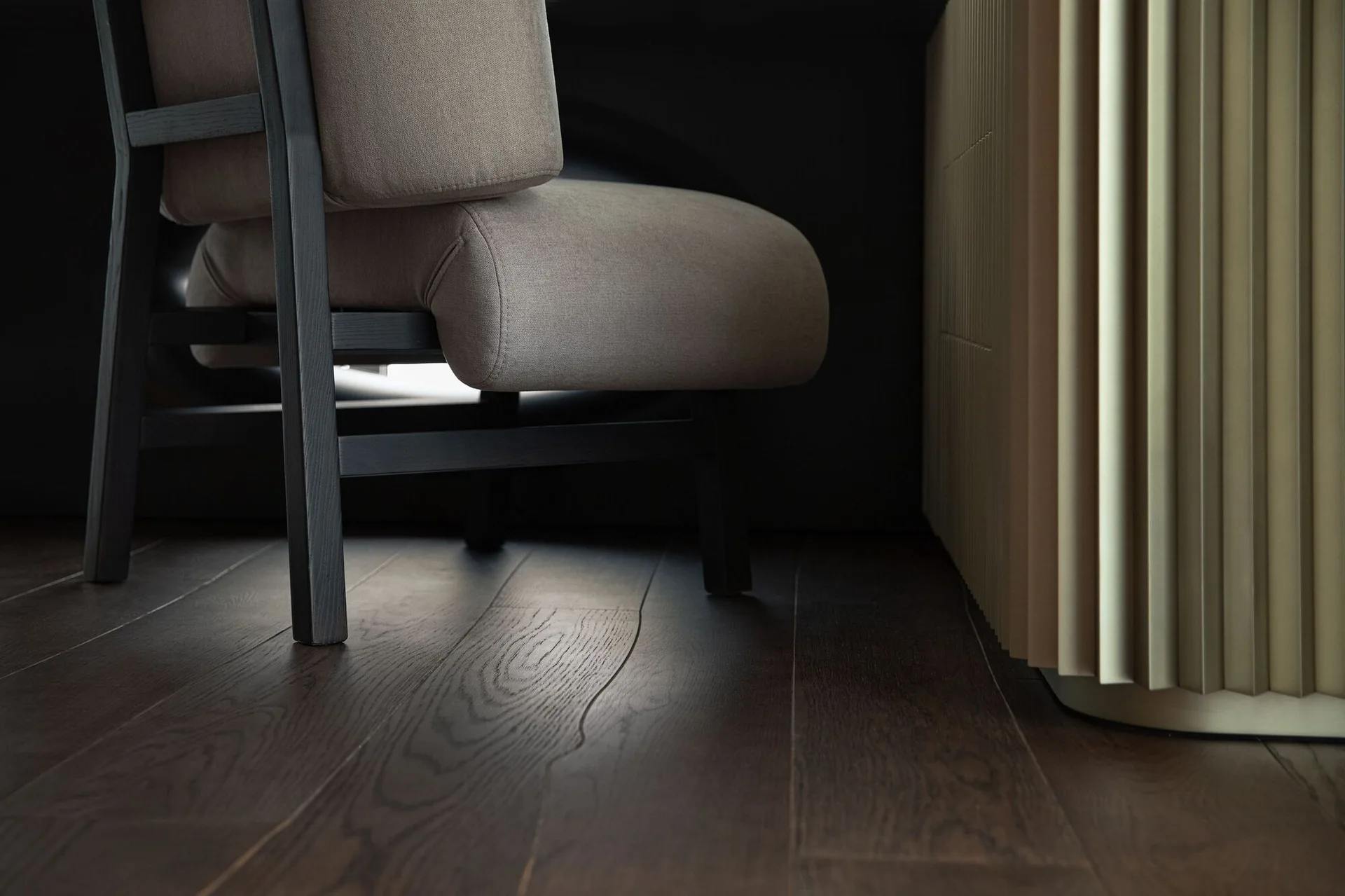 A close-up view of a beige upholstered armchair with a dark wood frame on a dark brown wood floor next to a vertically fluted wood panel.