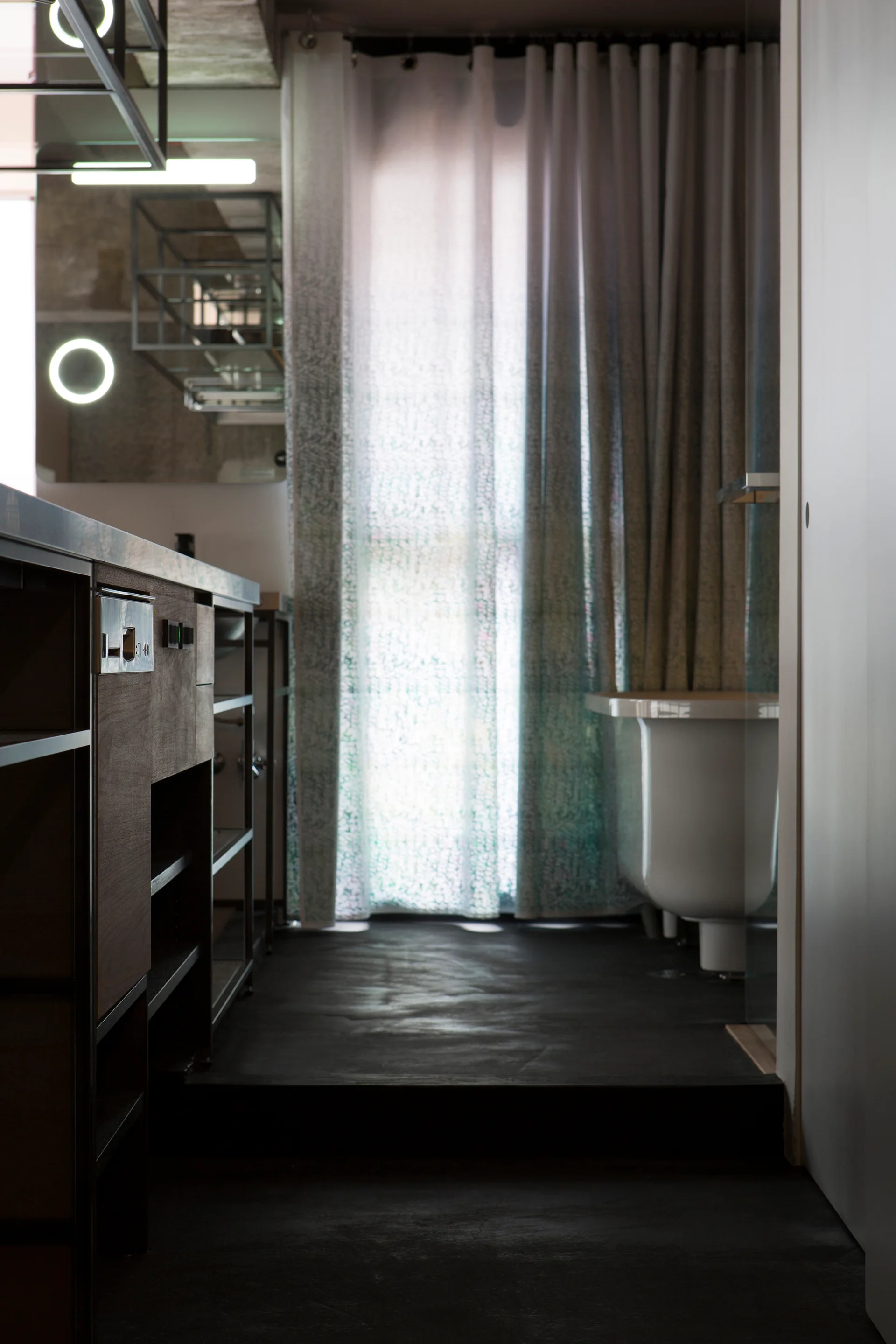 A modern industrial bathroom with dark concrete flooring, an open wood vanity, and a white freestanding bathtub positioned near sheer patterned curtains.