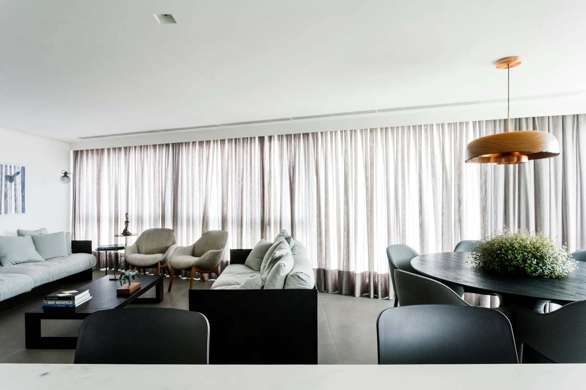 An open-concept living and dining area features a grey sofa, curved armchairs, a round dining table, and floor-to-ceiling sheer grey curtains.