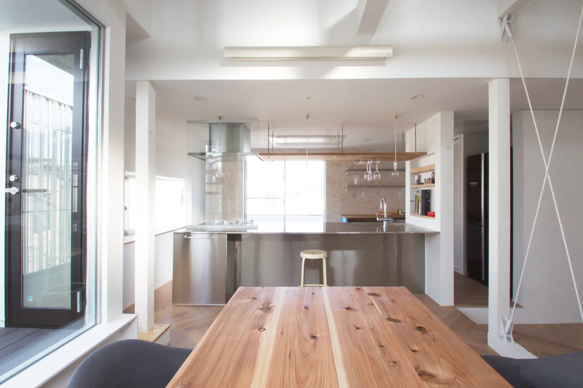 A modern kitchen features a stainless steel island and a natural wood dining table in the foreground against white walls and herringbone floors.