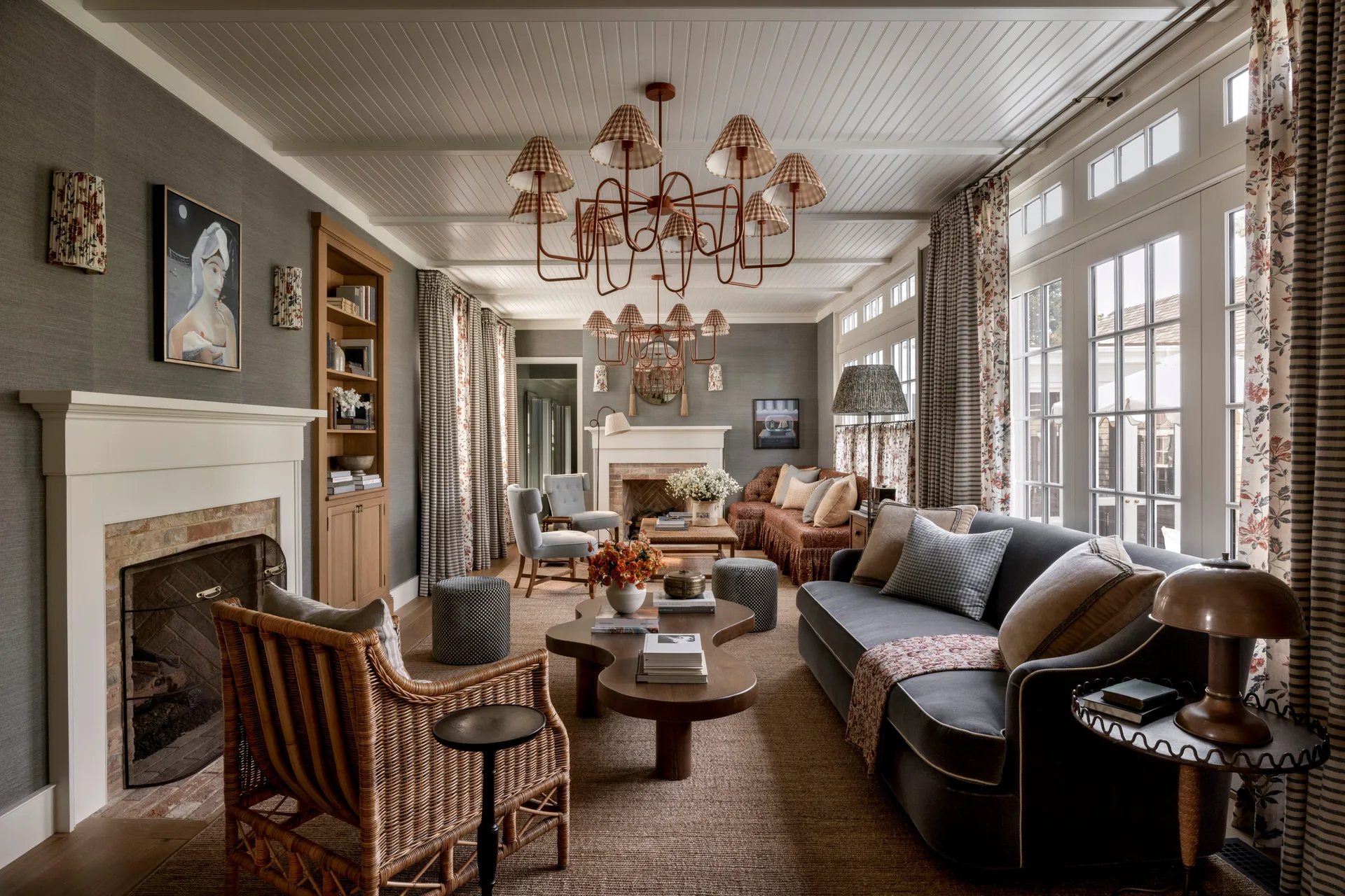 A traditional living room features a grey sofa, a wicker armchair, dual fireplaces, and a white planked ceiling with two tiered chandeliers.