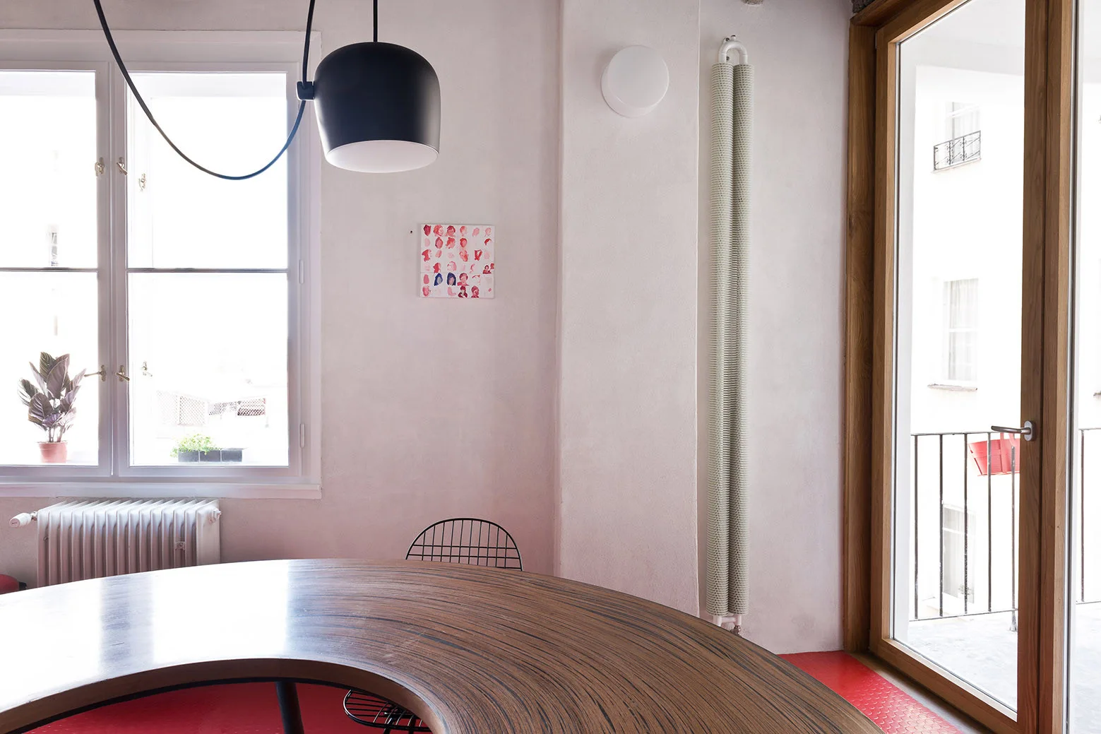 A minimalist dining room features a curved wooden table, a black pendant light, white walls, and a textured red floor next to a balcony door.