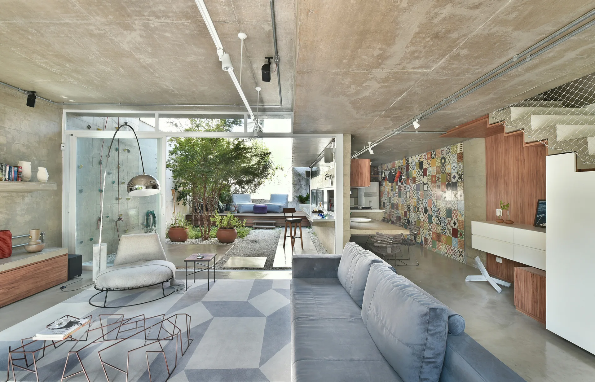 A modern industrial living room features concrete ceilings, a grey sofa, and sliding glass doors opening to a small patio with a climbing wall.