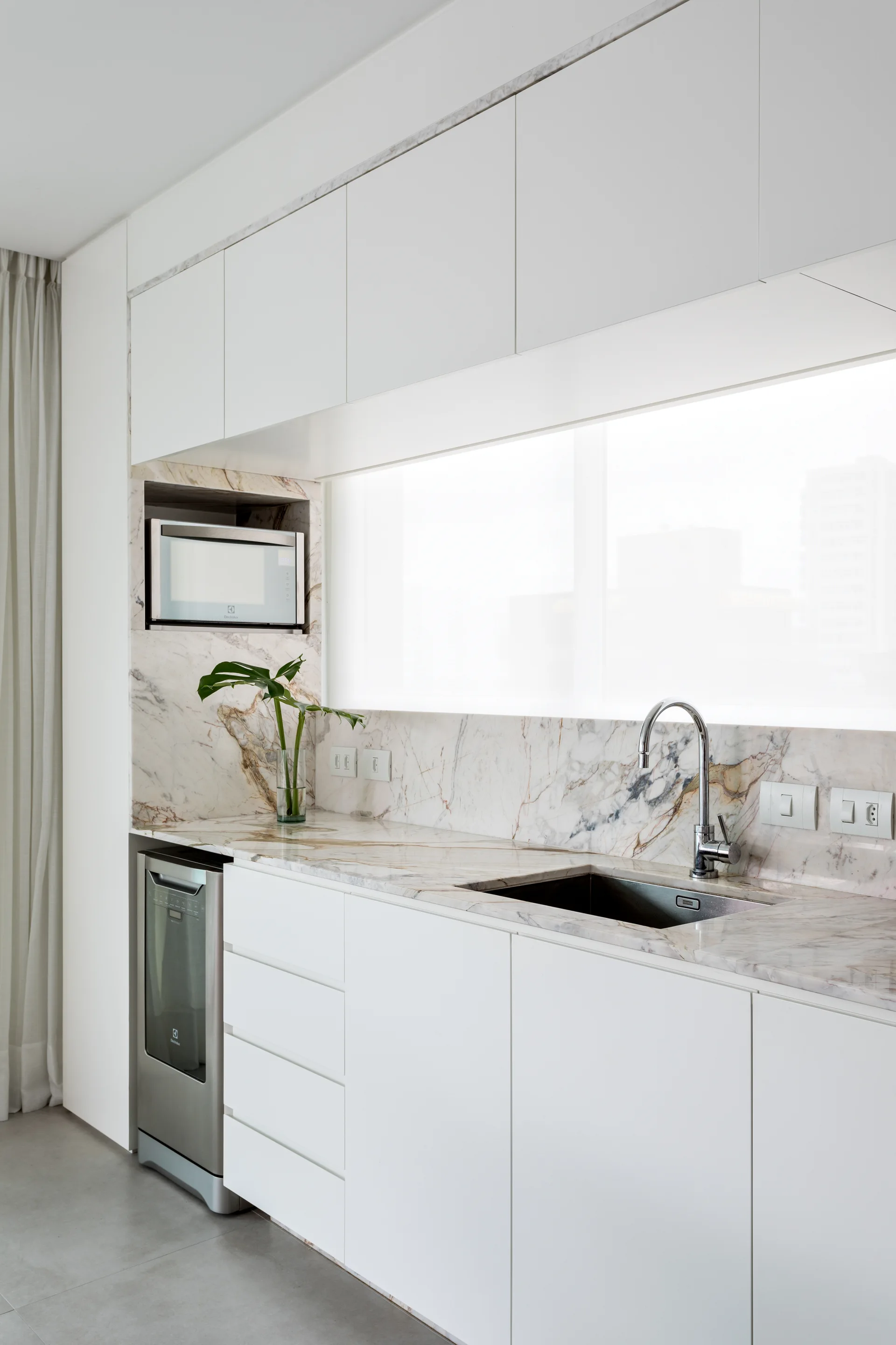 A minimalist kitchen features seamless white cabinets, a highly veined marble countertop, an integrated sink, and a built-in microwave.