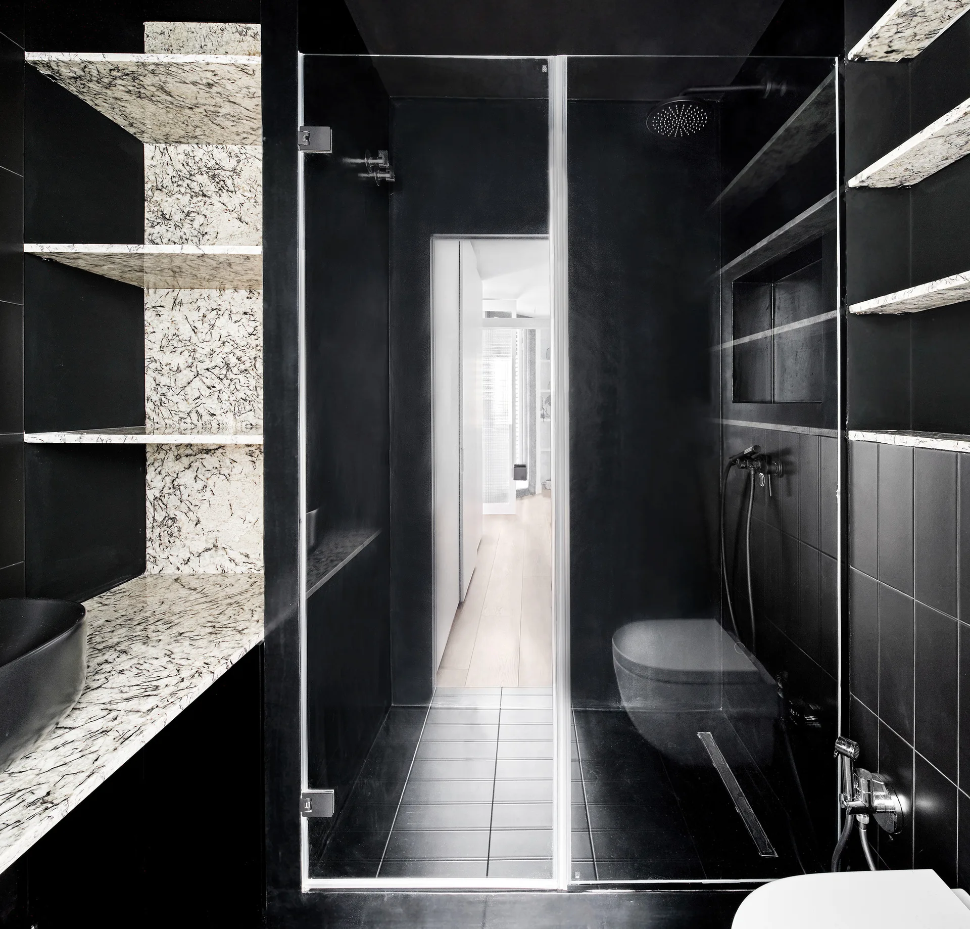 A modern black bathroom features a glass-enclosed shower, black square tiles, and built-in open shelves made of speckled white stone.