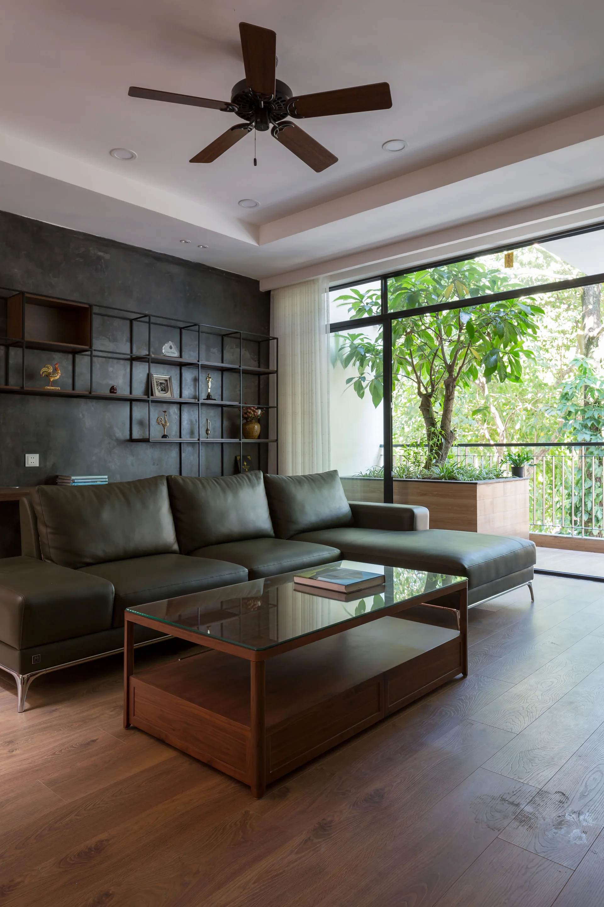 A modern living room features an olive green leather sectional sofa, a glass-top coffee table, and a dark textured wall next to a balcony.