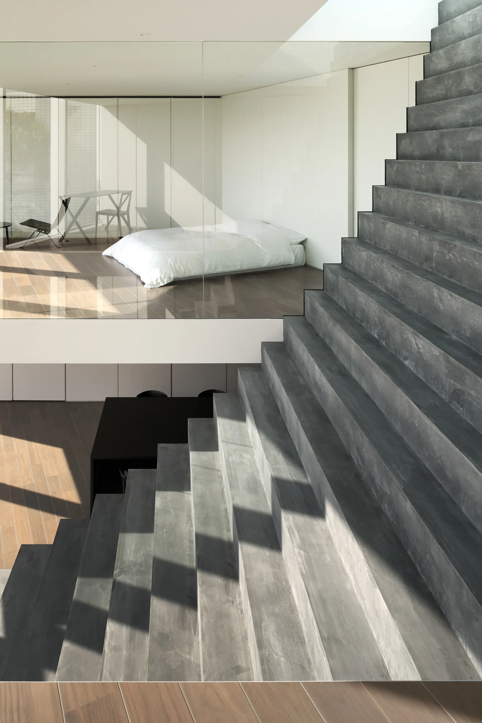 A modern interior featuring a gray concrete staircase leading up to a loft bedroom with glass walls, wood floors, and a simple white bed.