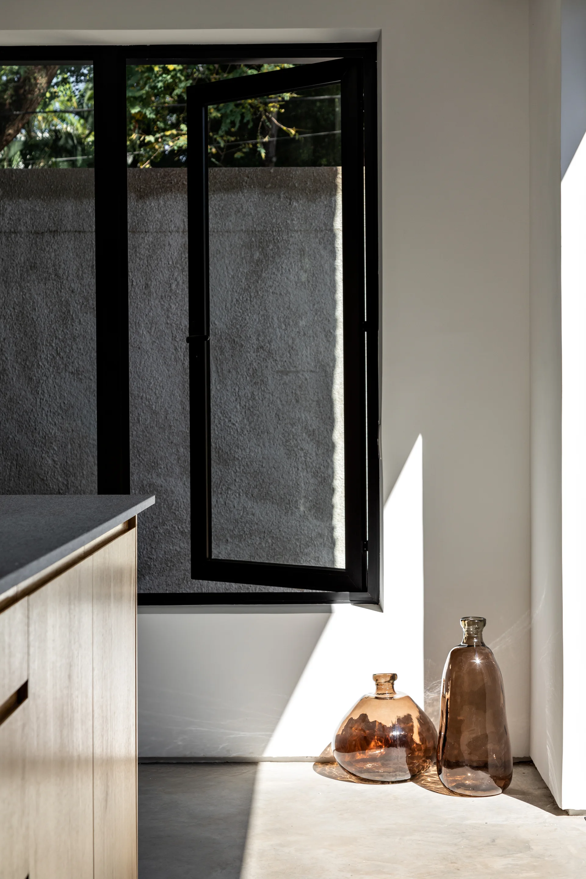 A minimalist kitchen corner features a black-framed window, light wood cabinetry, and two amber glass bottles on a concrete floor in bright sunlight.