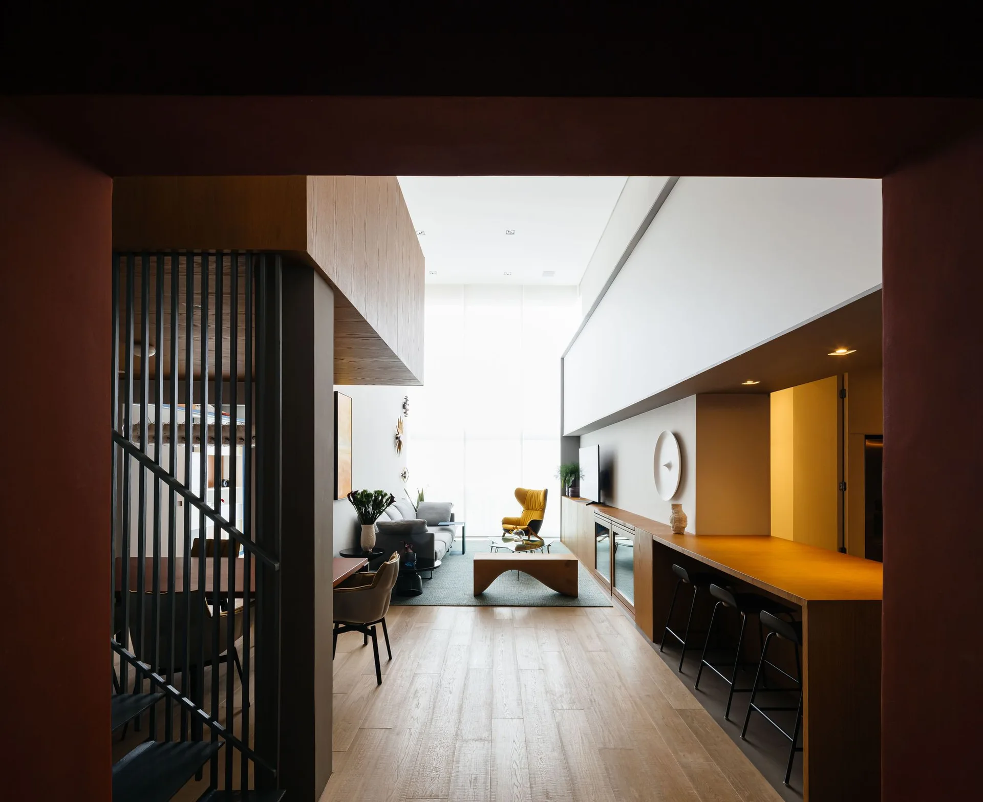 An open-plan living space features a long wood-topped bar with black stools, a staircase with metal railings, and a double-height ceiling.