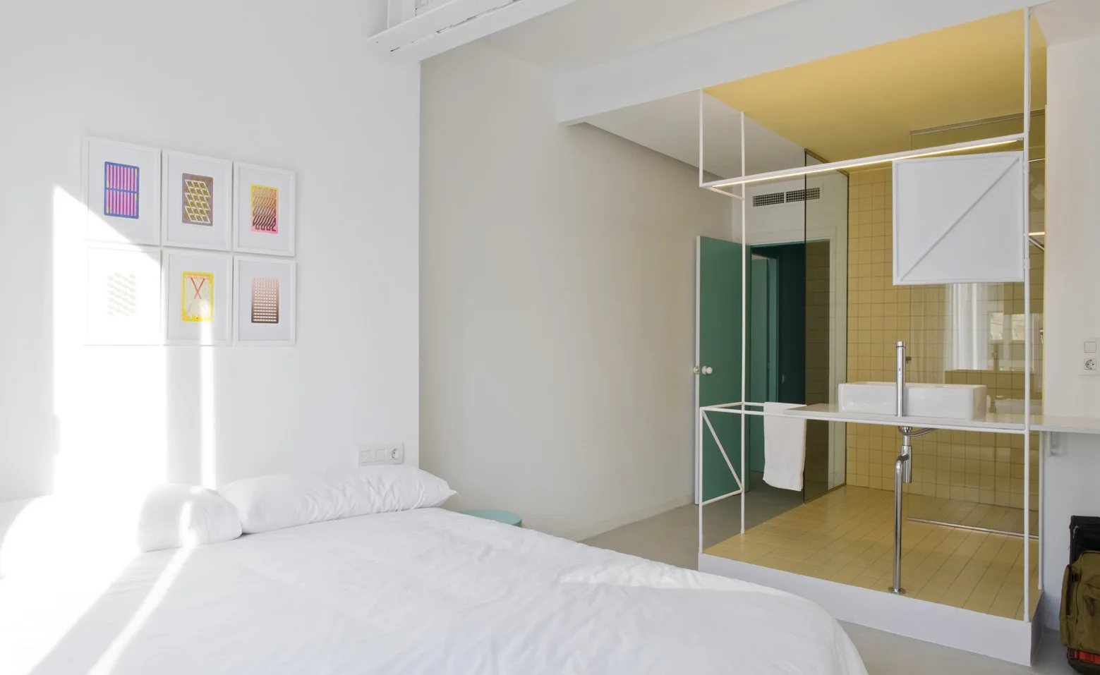 A minimalist white bedroom featuring a grid of artwork and an integrated bathroom area enclosed by a white metal frame and yellow tile.