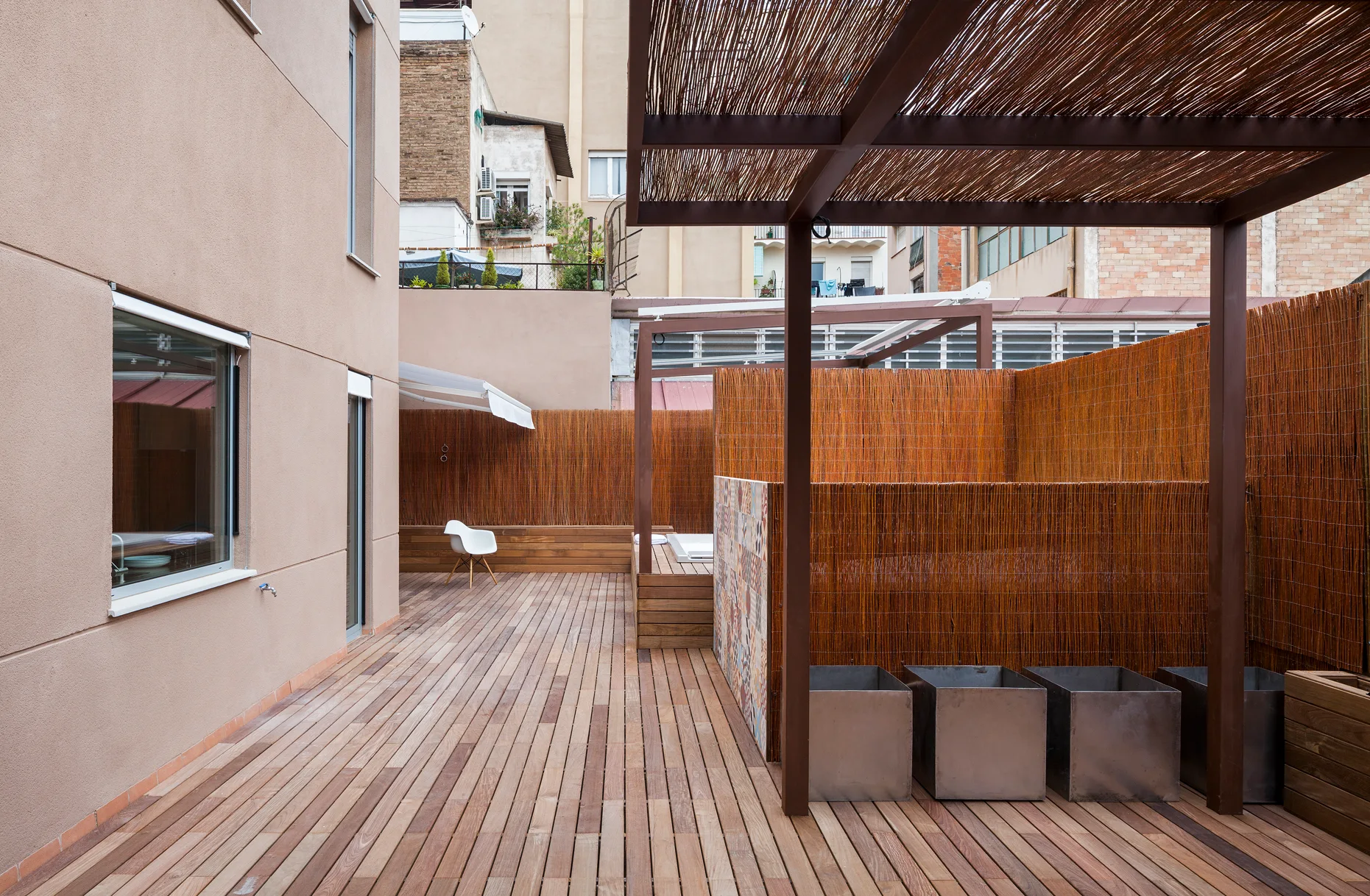 An urban rooftop terrace features a wooden plank deck, a brown metal pergola with a bamboo canopy, and high bamboo privacy fencing.