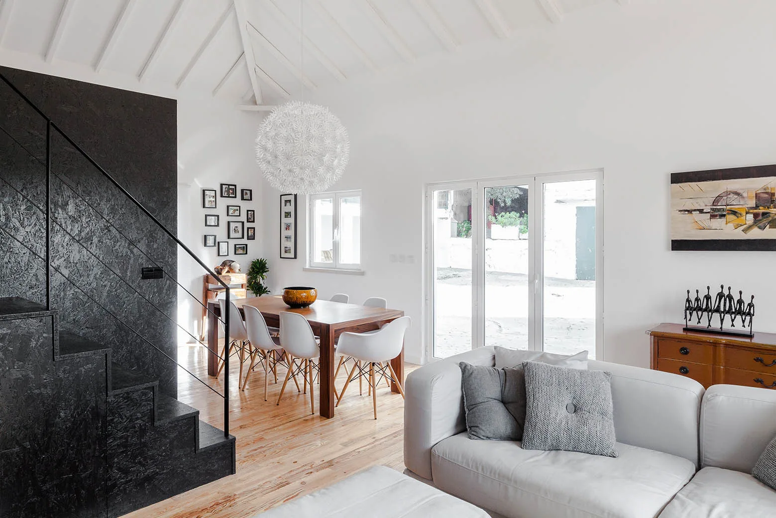 A bright open-concept living area features a black OSB staircase, a wooden dining table with white chairs, and a white vaulted ceiling.