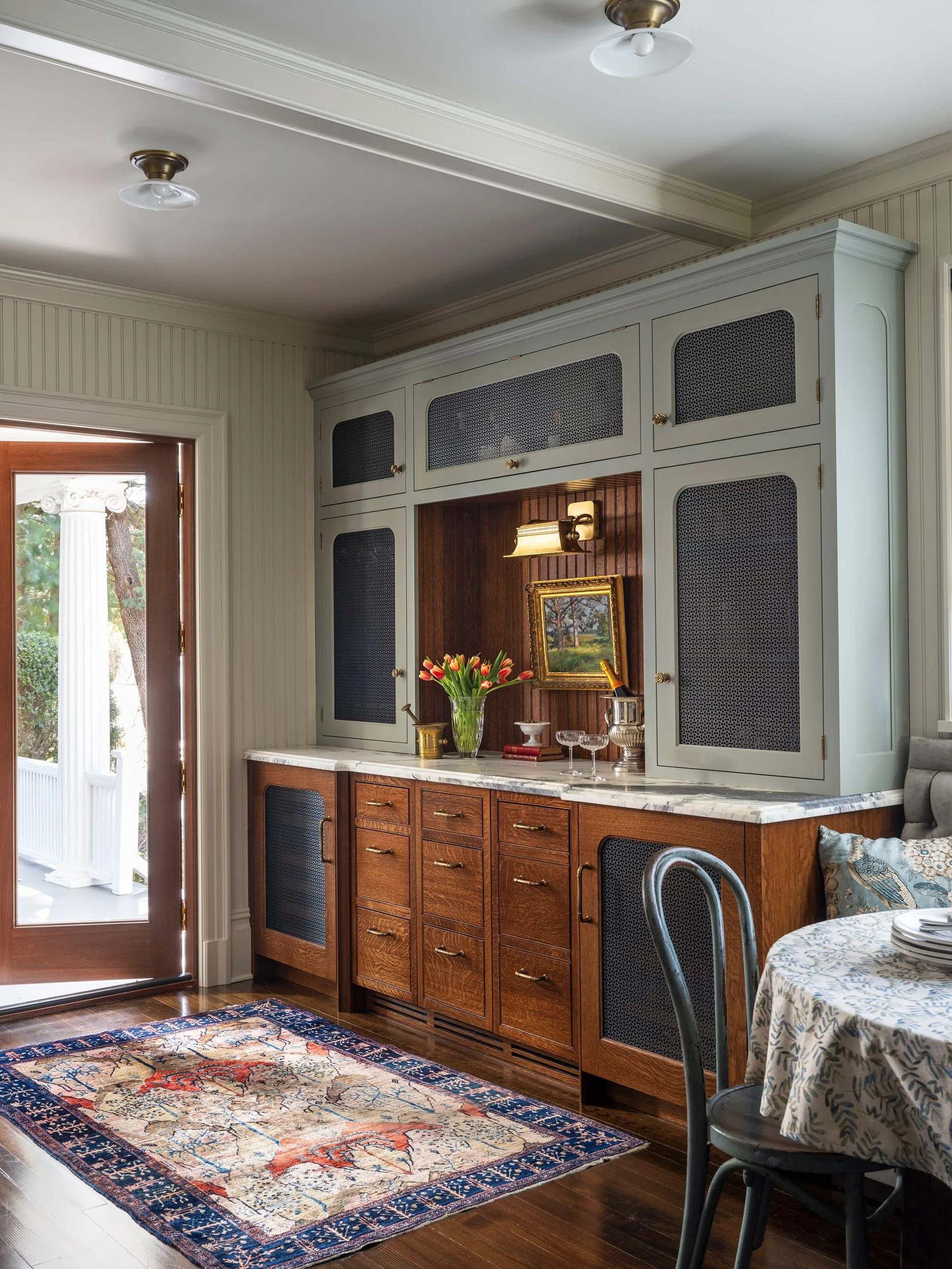 Built-in home bar with sage green upper cabinets, oak lower cabinets, marble countertops, and wire mesh door panels.