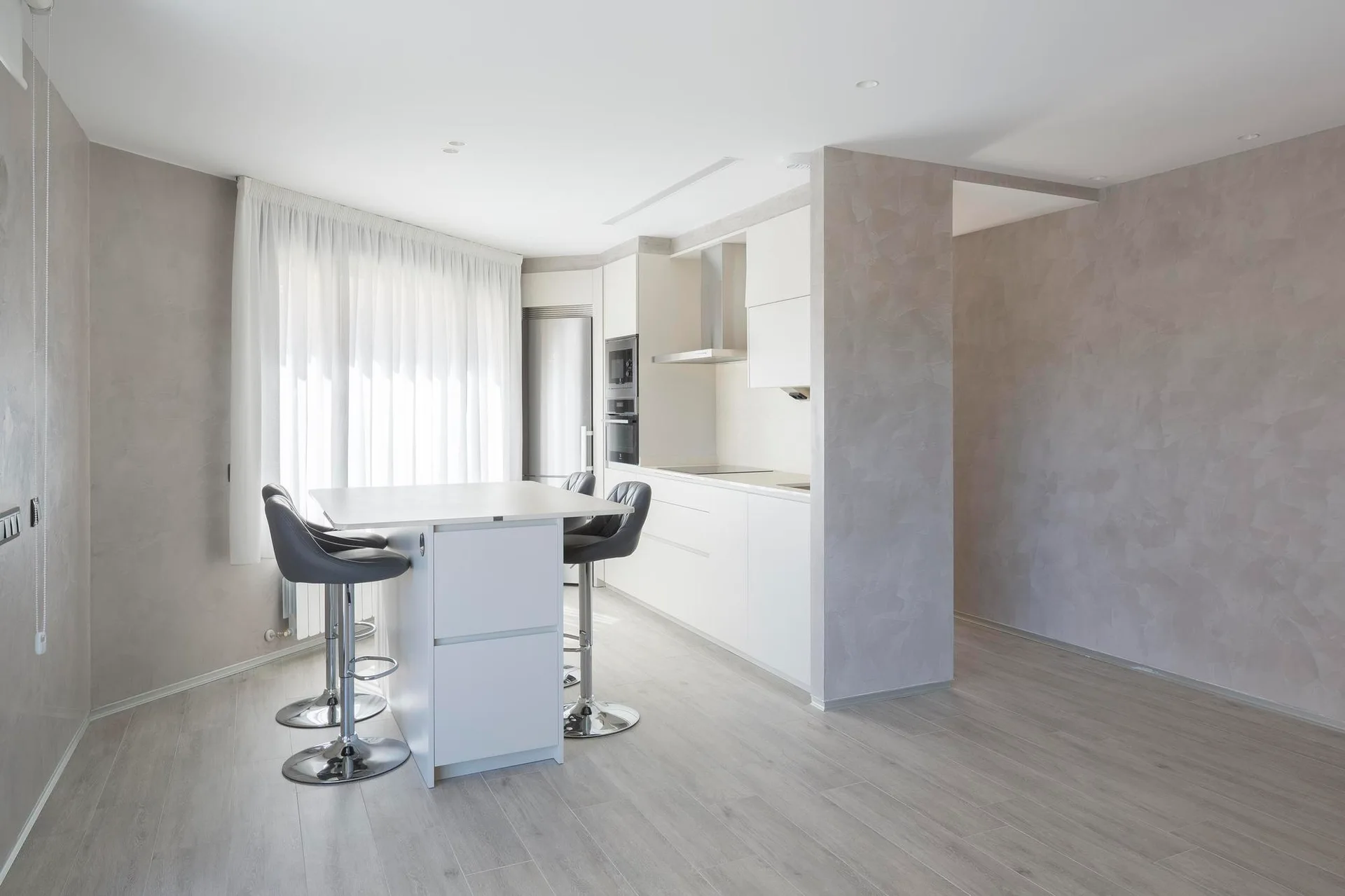 A contemporary kitchen features flat-front white cabinets, a matching island with grey bar stools, and textured grey plaster walls.