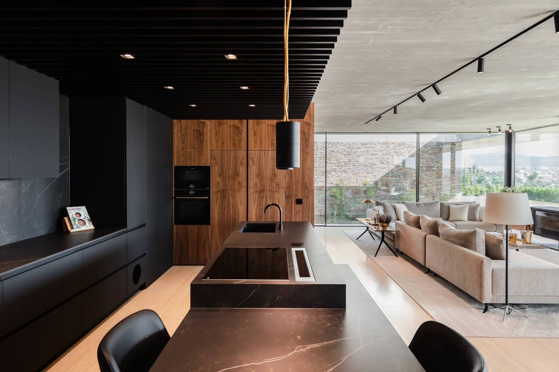 A modern kitchen features matte black cabinetry, a dark stone island, and a black slatted ceiling next to an open-concept living room.