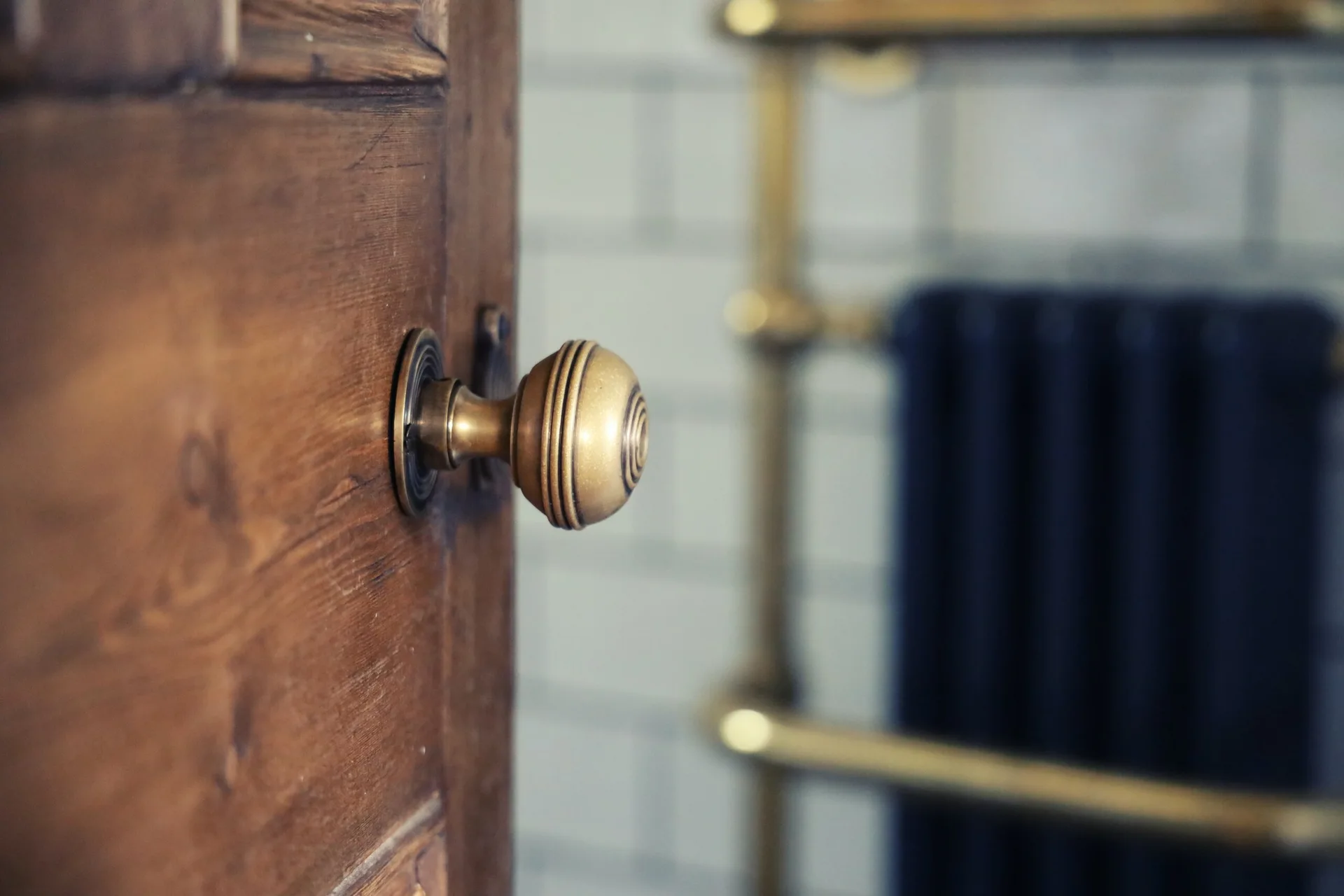 A close-up of a ribbed brass door knob on a stained wooden door, with blurred white subway tiles and a brass towel rack in the background.