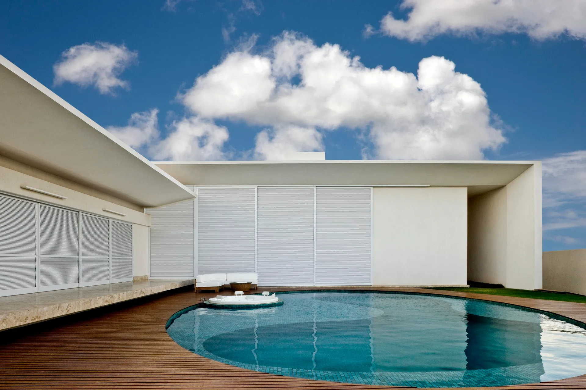 A circular blue tiled swimming pool is set into a warm wood plank deck next to a modern white building with louvered sliding panels.