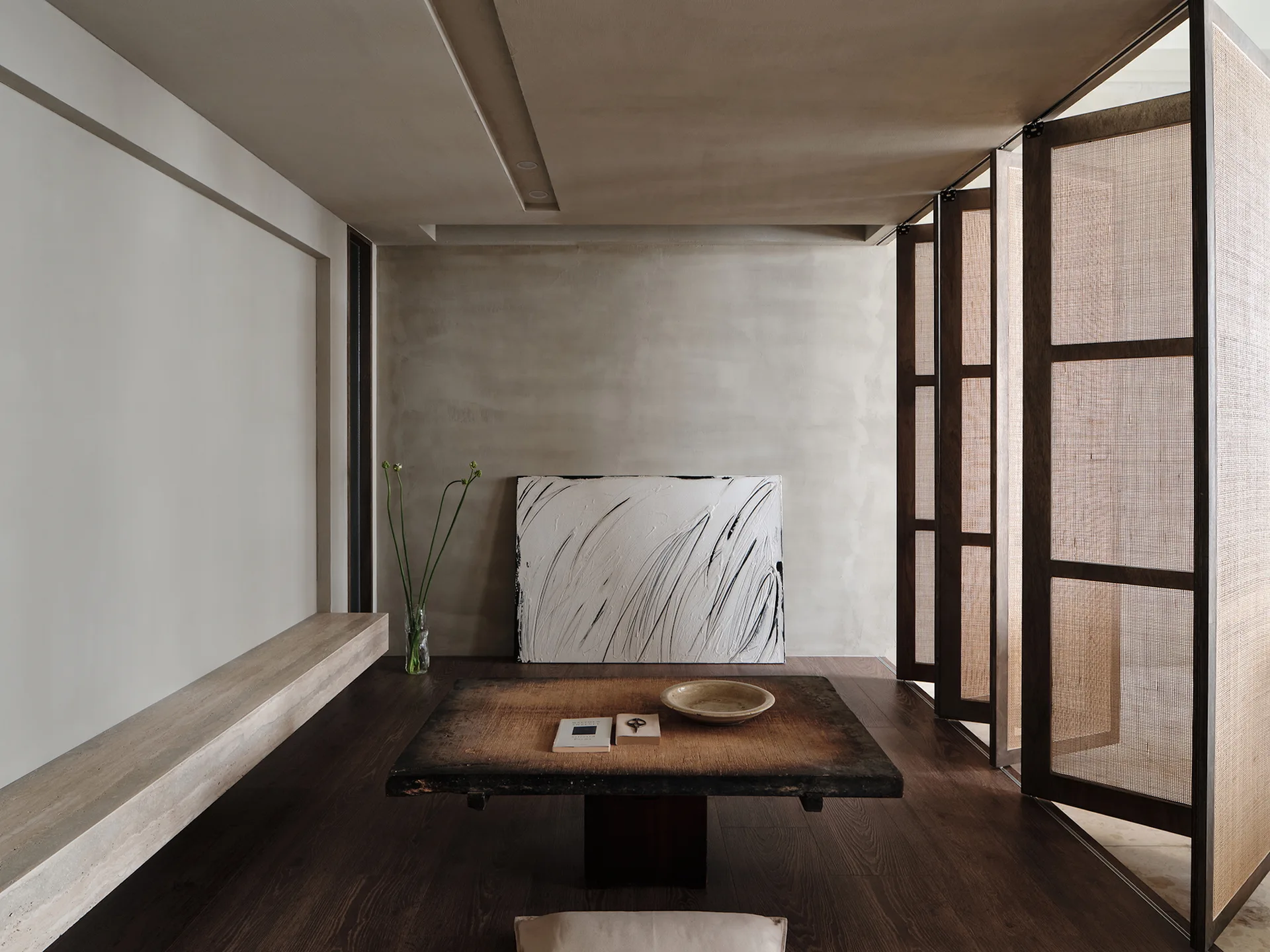 A minimalist room features a low wood coffee table, a floating stone shelf, abstract art resting on the floor, and tall woven screen panels.