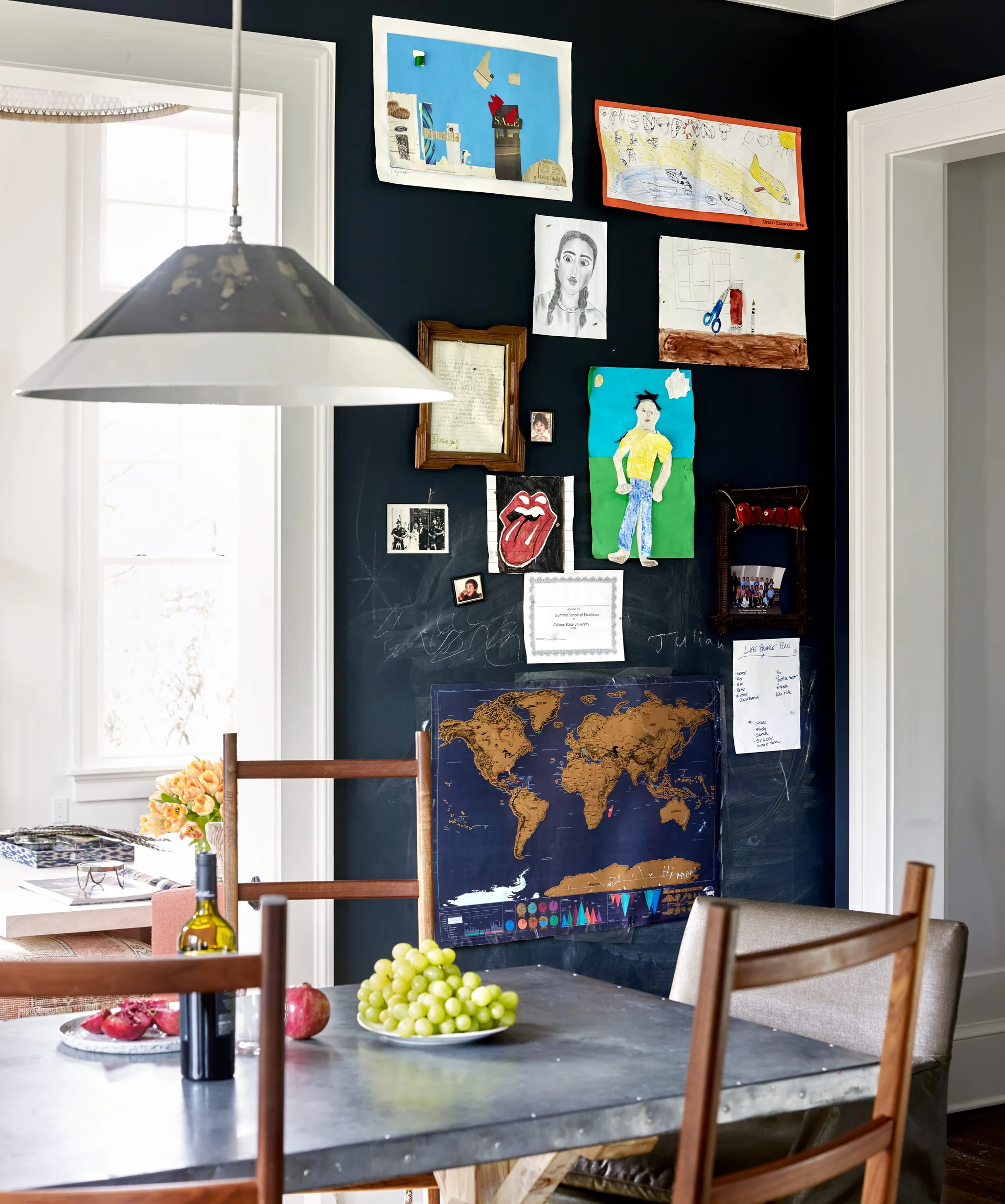 A metal dining table and wooden chairs stand before a dark chalkboard wall decorated with children's colorful artwork, sketches, and a map.