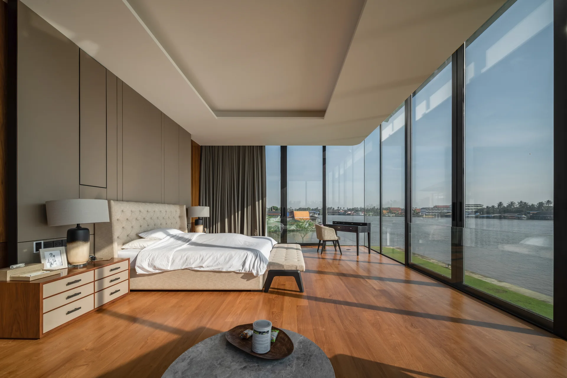A modern bedroom features a tufted beige bed, warm wood flooring, and floor-to-ceiling glass walls overlooking a wide river under a blue sky.