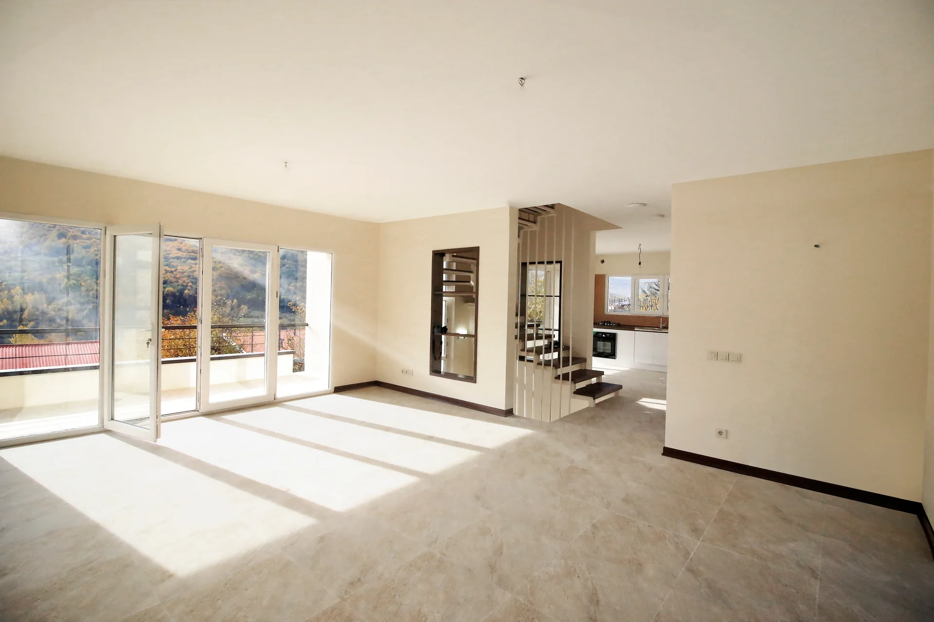 An empty beige living room featuring large-format floor tiles, a minimalist open staircase, and wide glass doors leading to a sunny balcony.