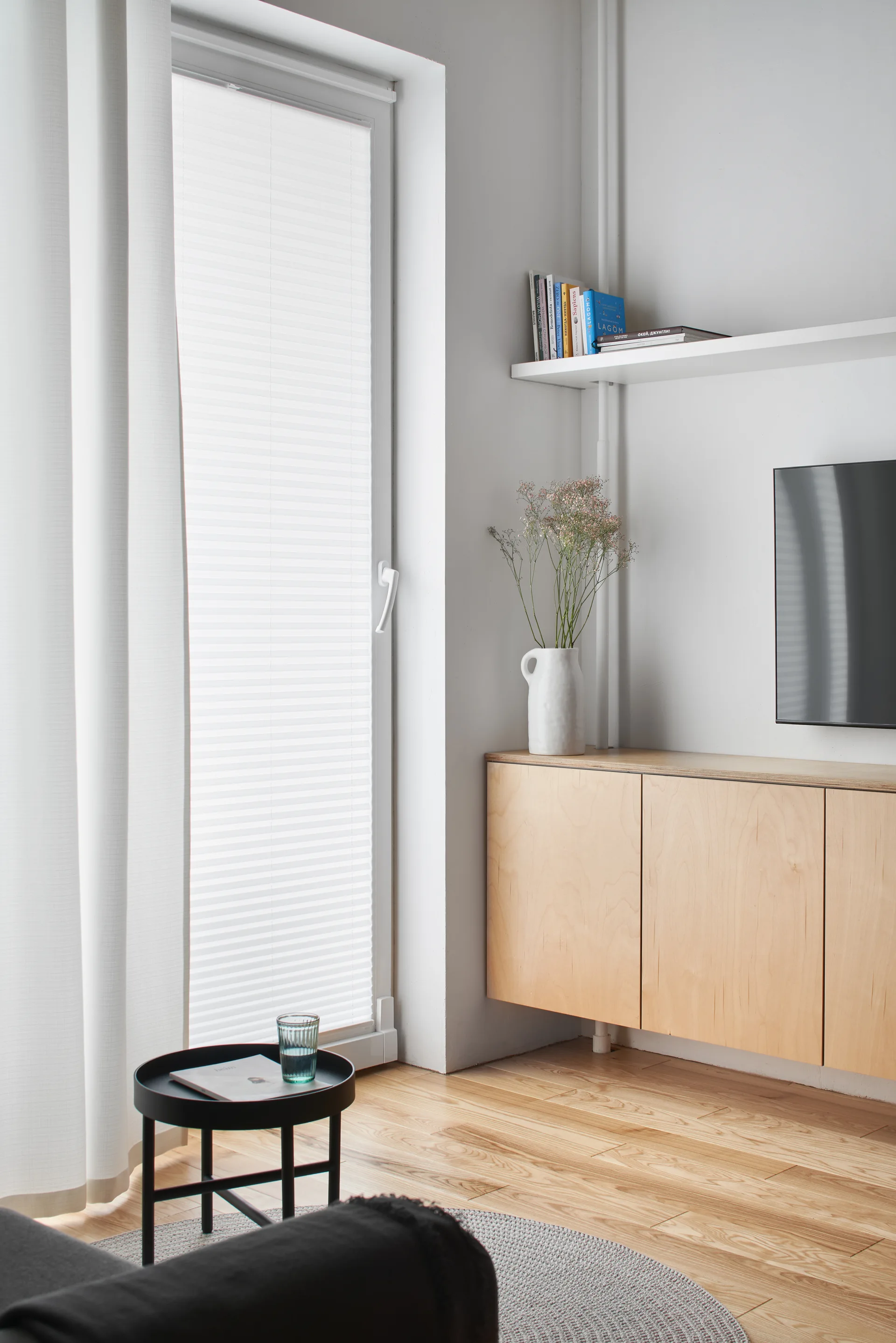A minimalist living area features a wall-mounted light wood cabinet, a white floating shelf with books, and a small round black side table.