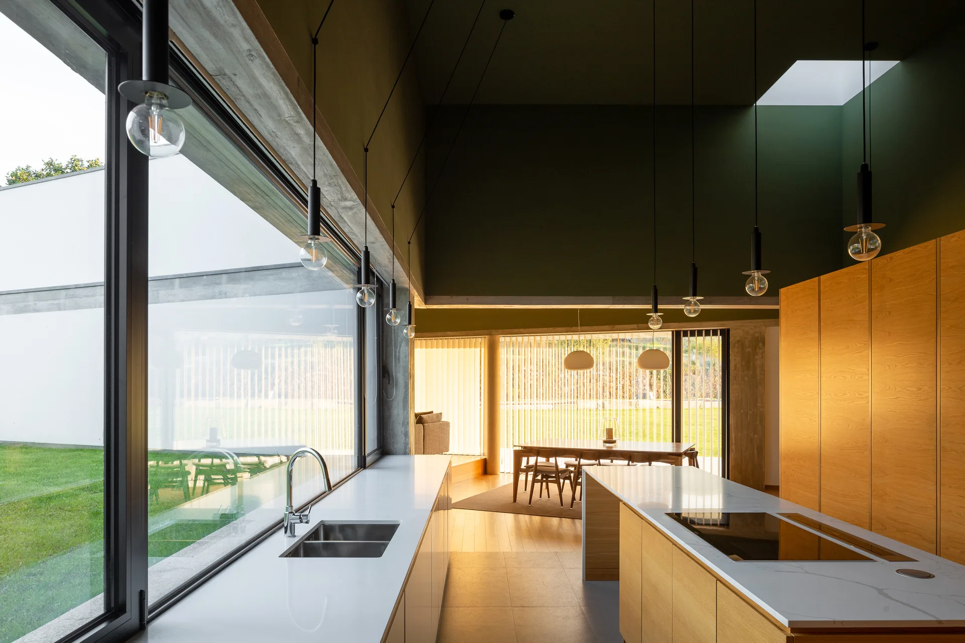 A minimalist kitchen features a forest green ceiling with a skylight, light wood cabinets, concrete structural details, and a large window.