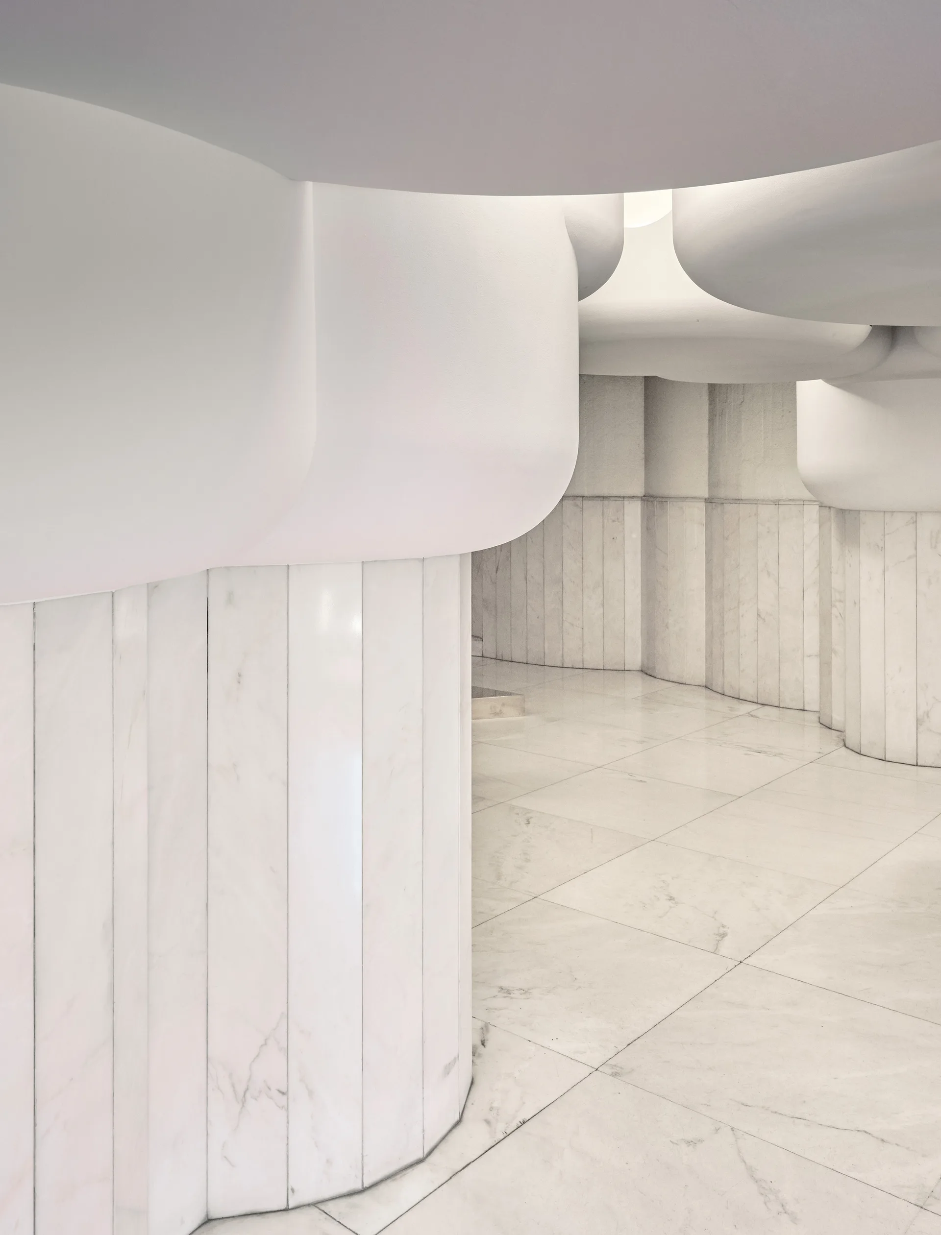 A minimalist white interior features organic sculptural ceiling forms suspended above fluted marble wall columns and a matching polished marble tile floor.