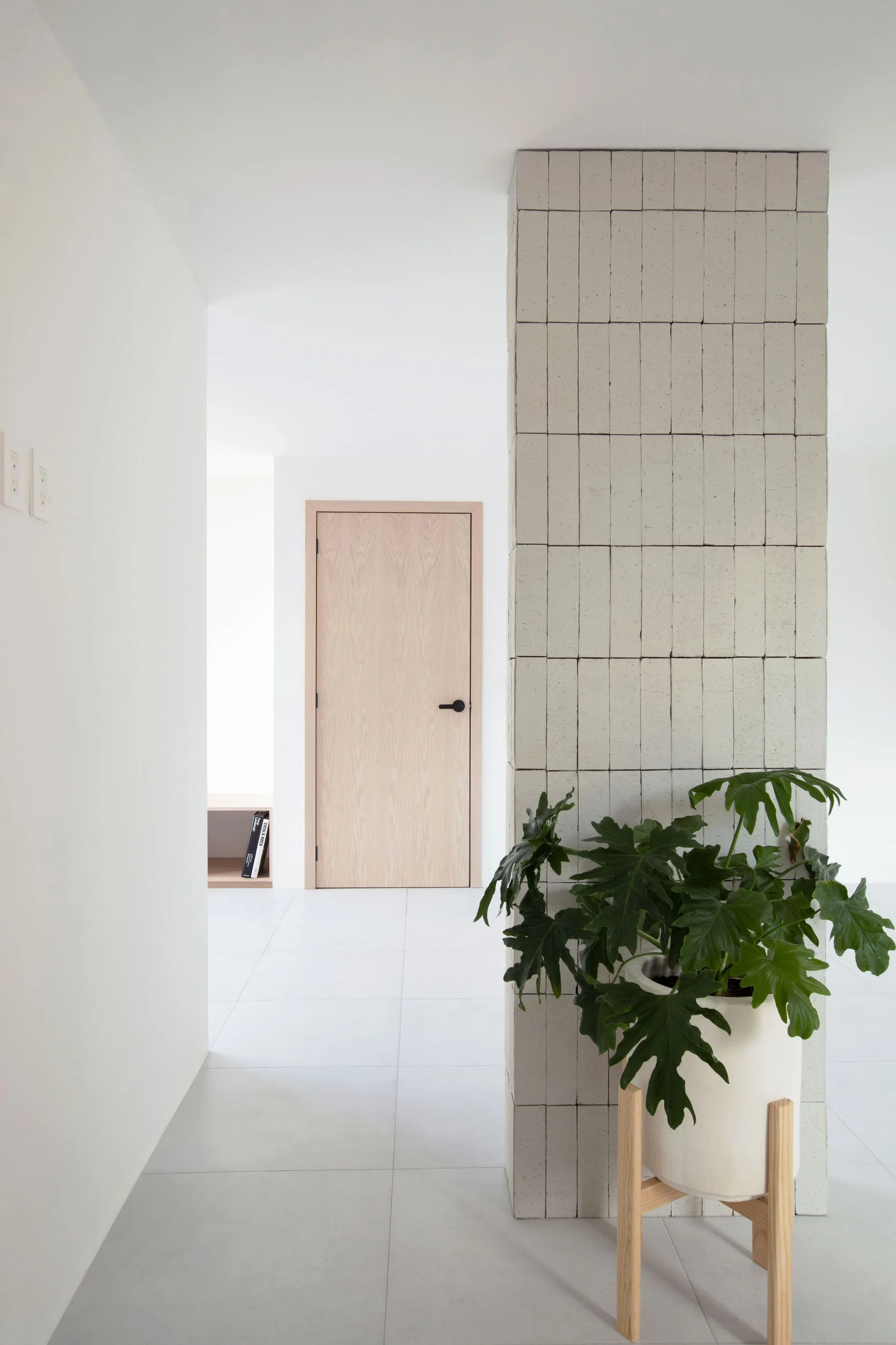 A minimalist white hallway features a structural column clad in stacked light gray tiles next to a green potted plant and a light wood door.
