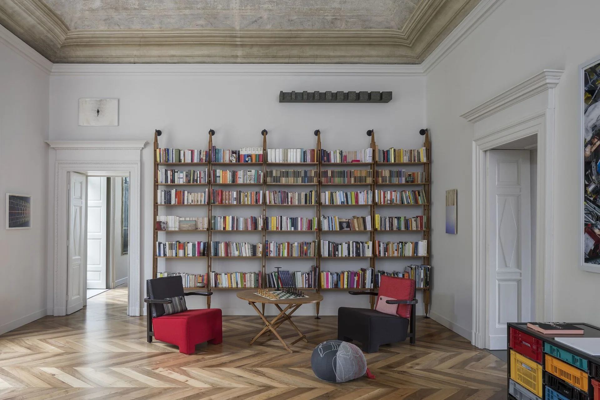 A high-ceilinged room features a large wall-spanning wooden bookshelf, two modern armchairs, a round table with a chess set, and herringbone wood floors.