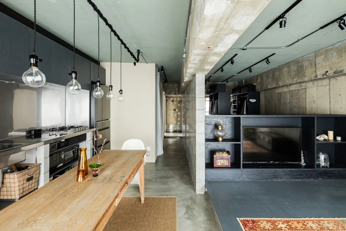 An industrial dining and kitchen area features a long wooden table, a row of exposed bulb pendants, and a painted sage green concrete ceiling.