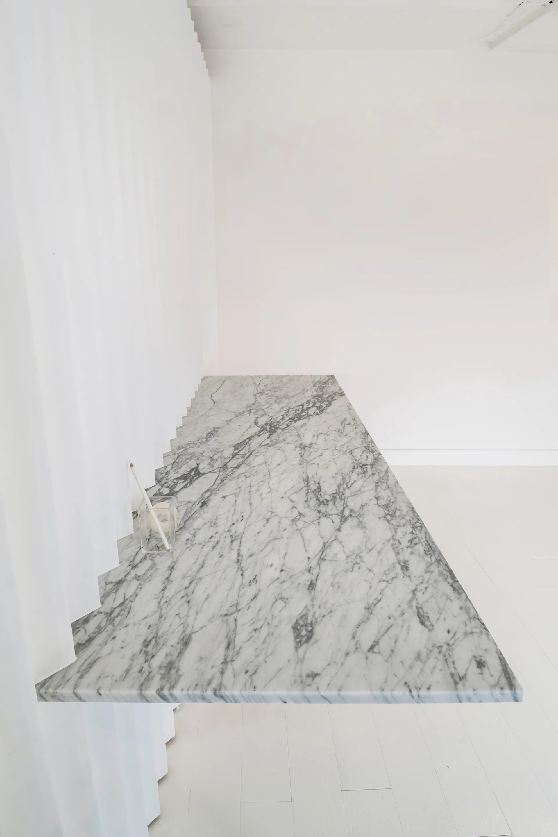A floating white marble countertop with grey veining extends next to a pleated white curtain in a minimalist white room with painted wood floors.