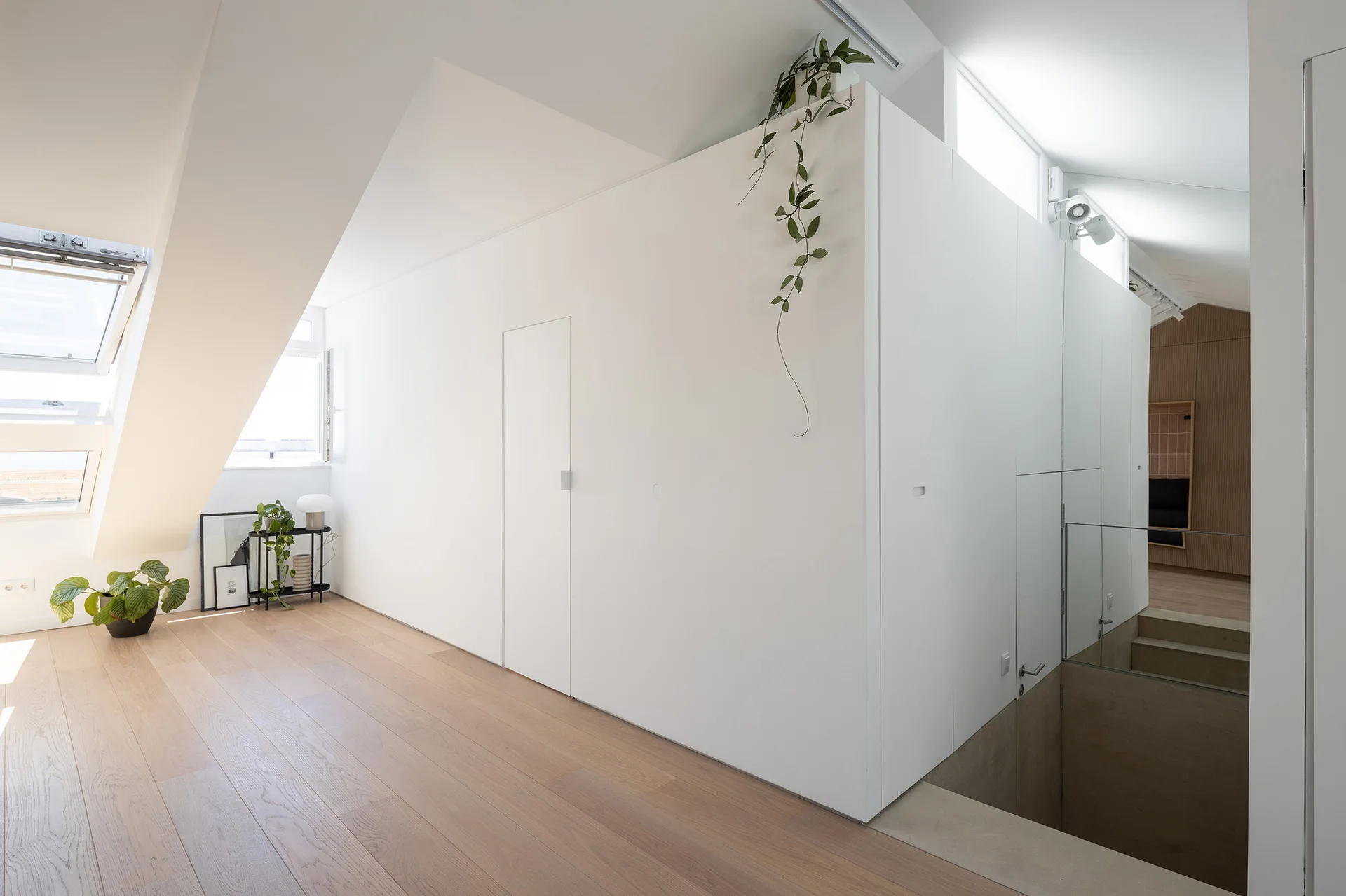A bright attic landing features a large white partition wall, light wood flooring, a sloped ceiling with a skylight, and stairs with a glass railing.