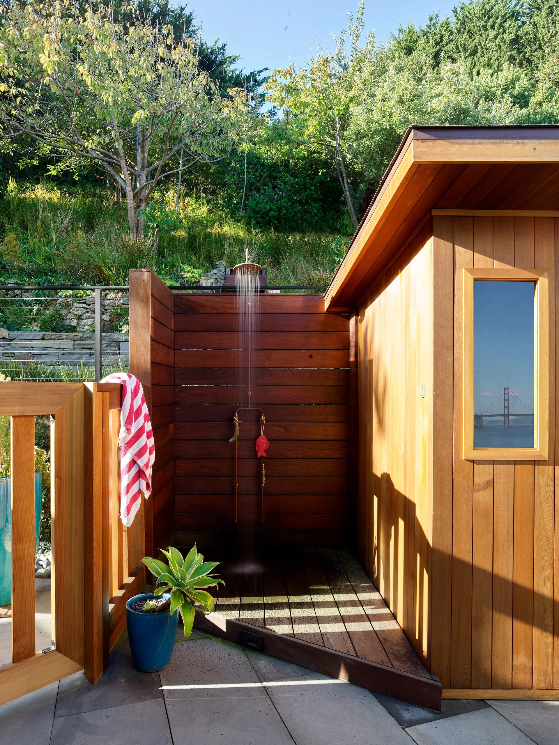 An outdoor shower built with stained wood planks features a brass showerhead, a blue potted plant, and a red and white striped towel.