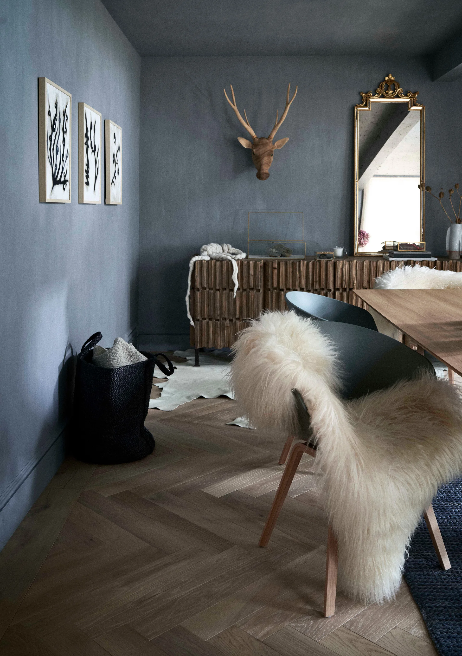 A dining room with charcoal gray walls features wooden dining chairs draped in white sheepskin, a herringbone wood floor, and a fluted wood sideboard.