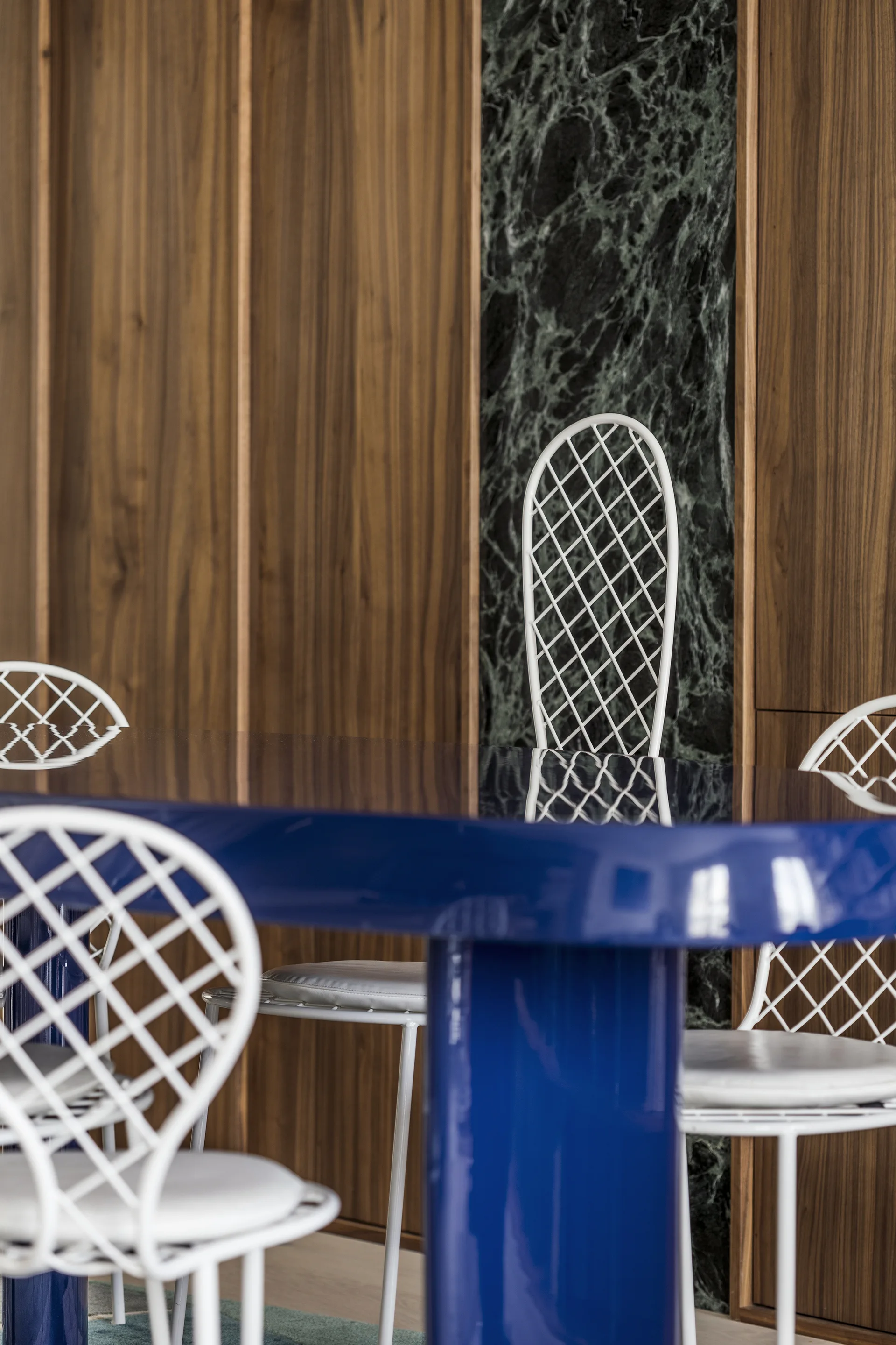 A glossy cobalt blue dining table is paired with white lattice back chairs against a background of wood wall paneling and a green marble insert.