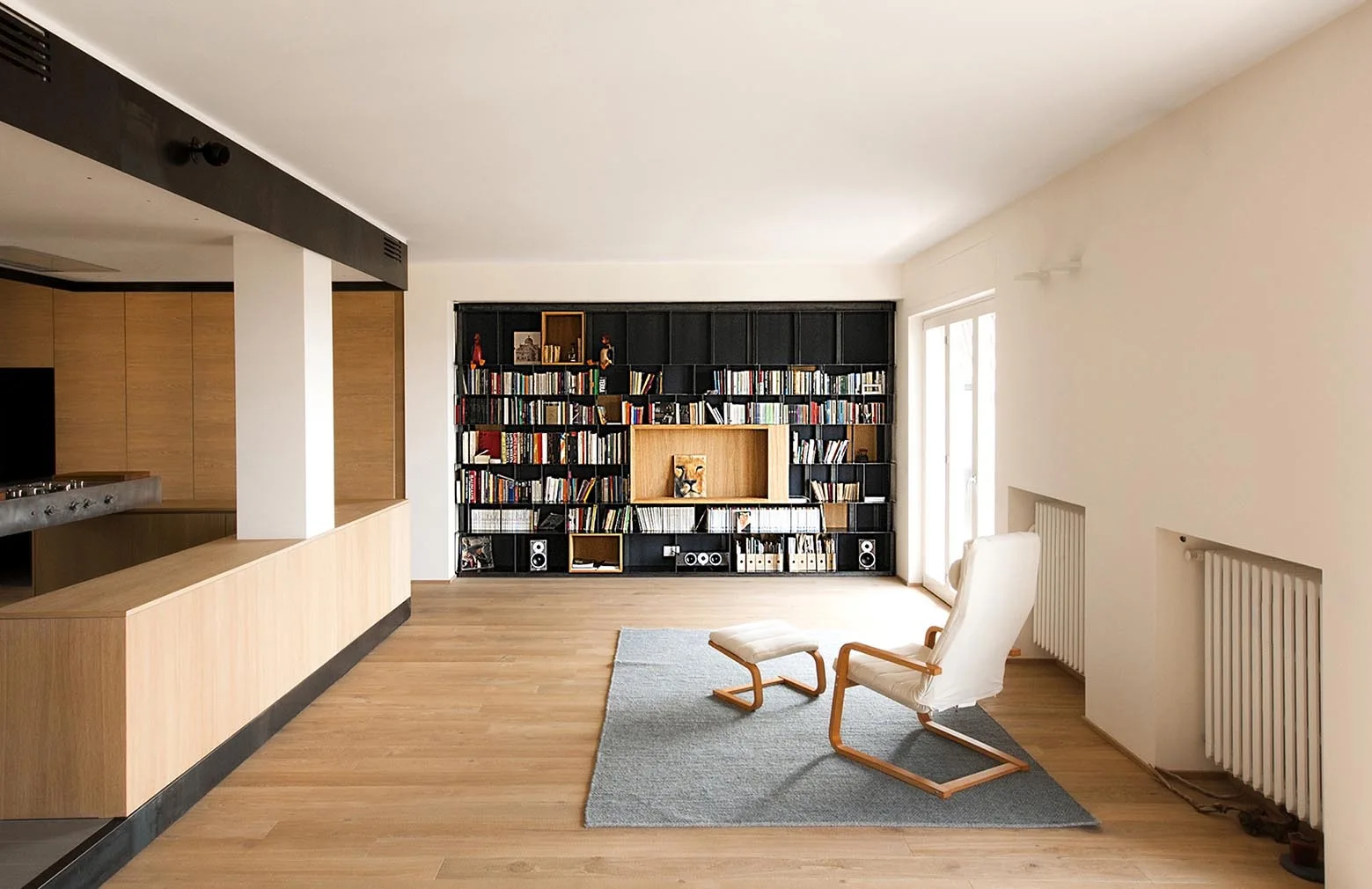 A minimalist living room features a large black wall-to-wall bookshelf, a white lounge chair with an ottoman on a grey rug, and light wood flooring.