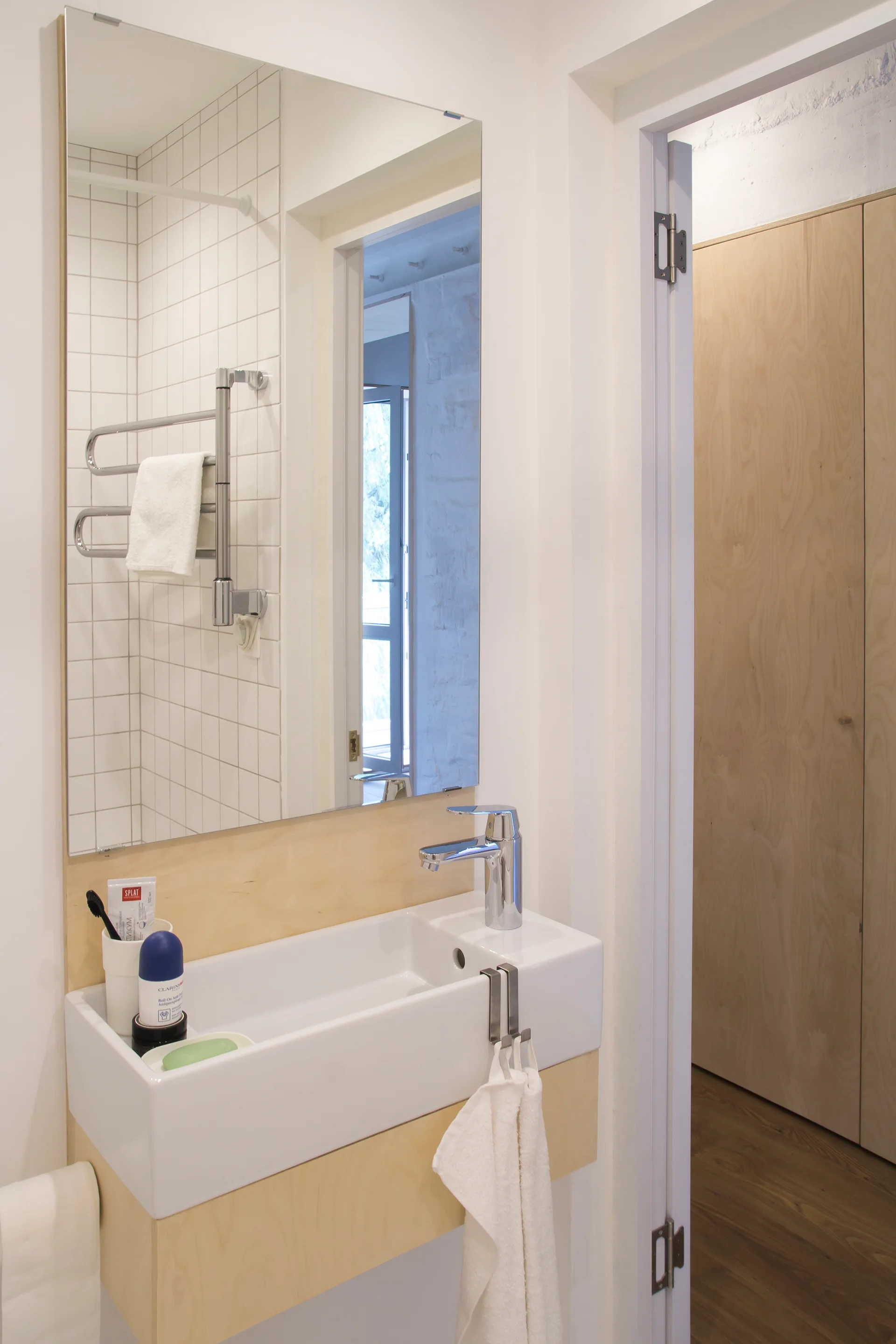 A minimalist bathroom features a wall-mounted plywood vanity with a rectangular white sink, a large mirror, and square white wall tiles.