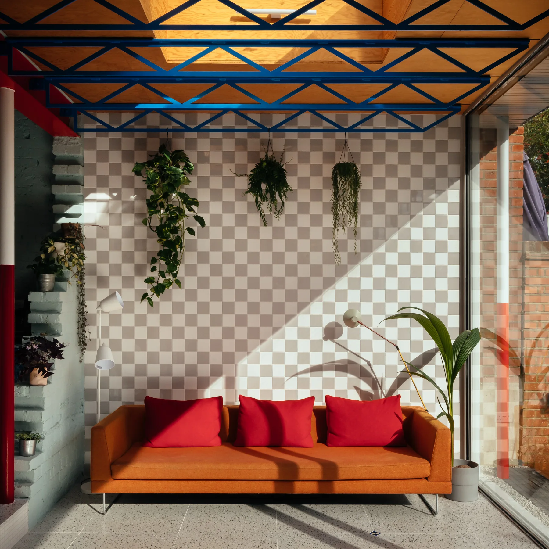 An orange sofa with red pillows sits against a grey and white checkerboard tiled wall under a blue metal roof truss.
