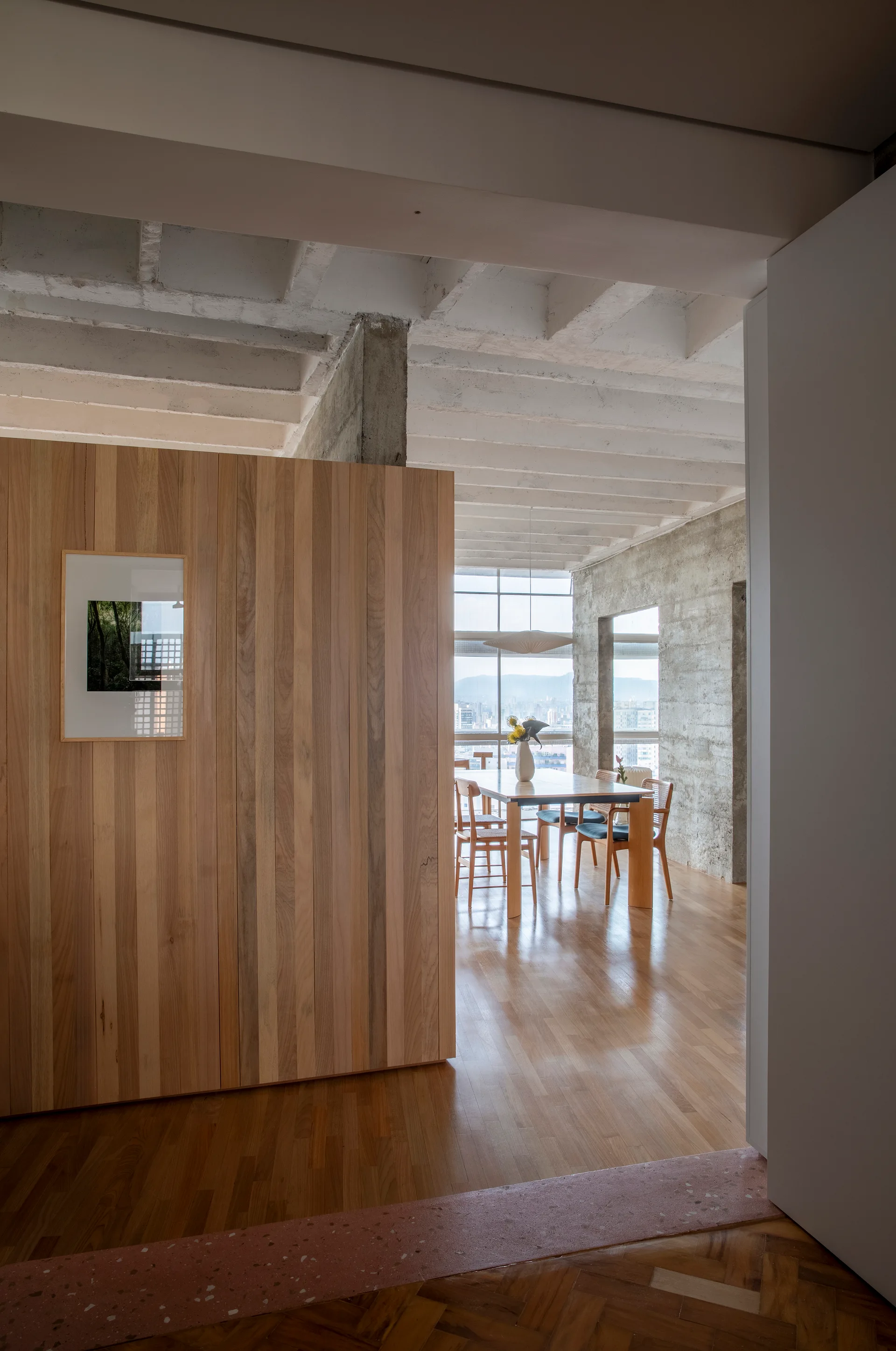 An open dining area features a light wood dining table, exposed concrete walls and ceiling beams, and a vertical wood-slat partition.