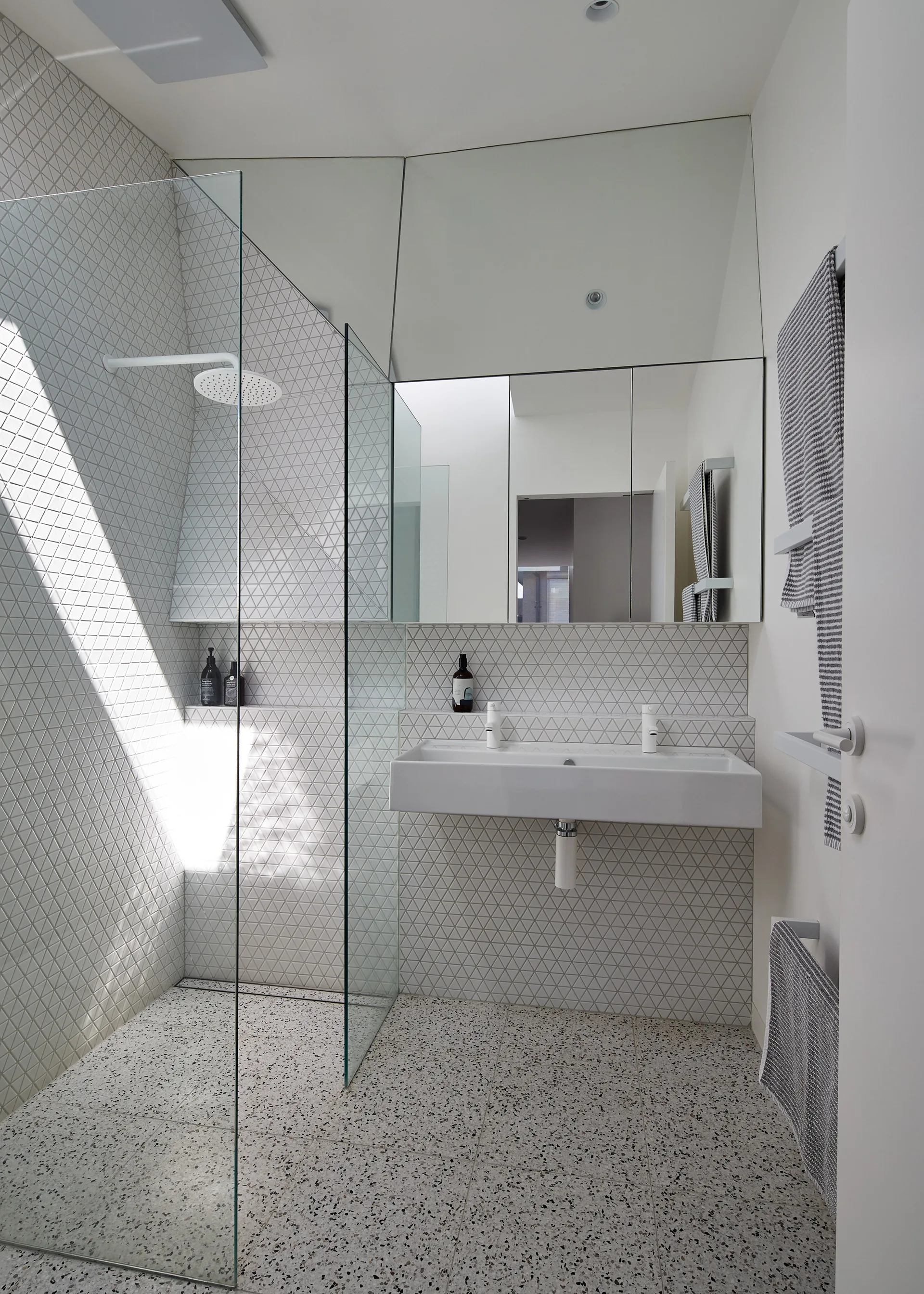 A contemporary bathroom features white geometric triangle wall tiles, a frameless glass shower screen, and a wall-mounted sink over speckled terrazzo flooring.