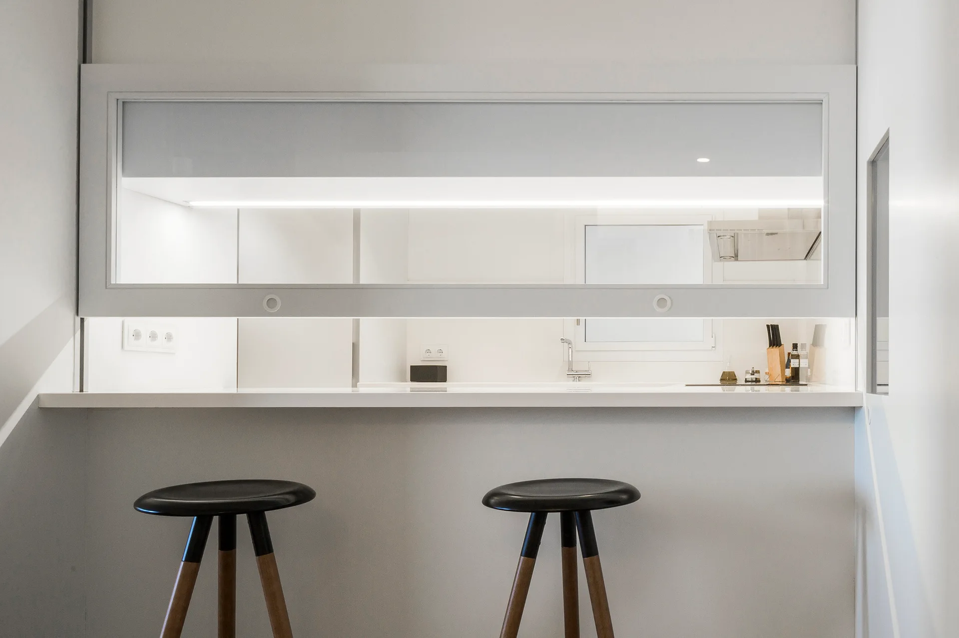A minimalist white breakfast bar features two black wood-legged stools beneath a large framed glass pass-through window looking into a kitchen.